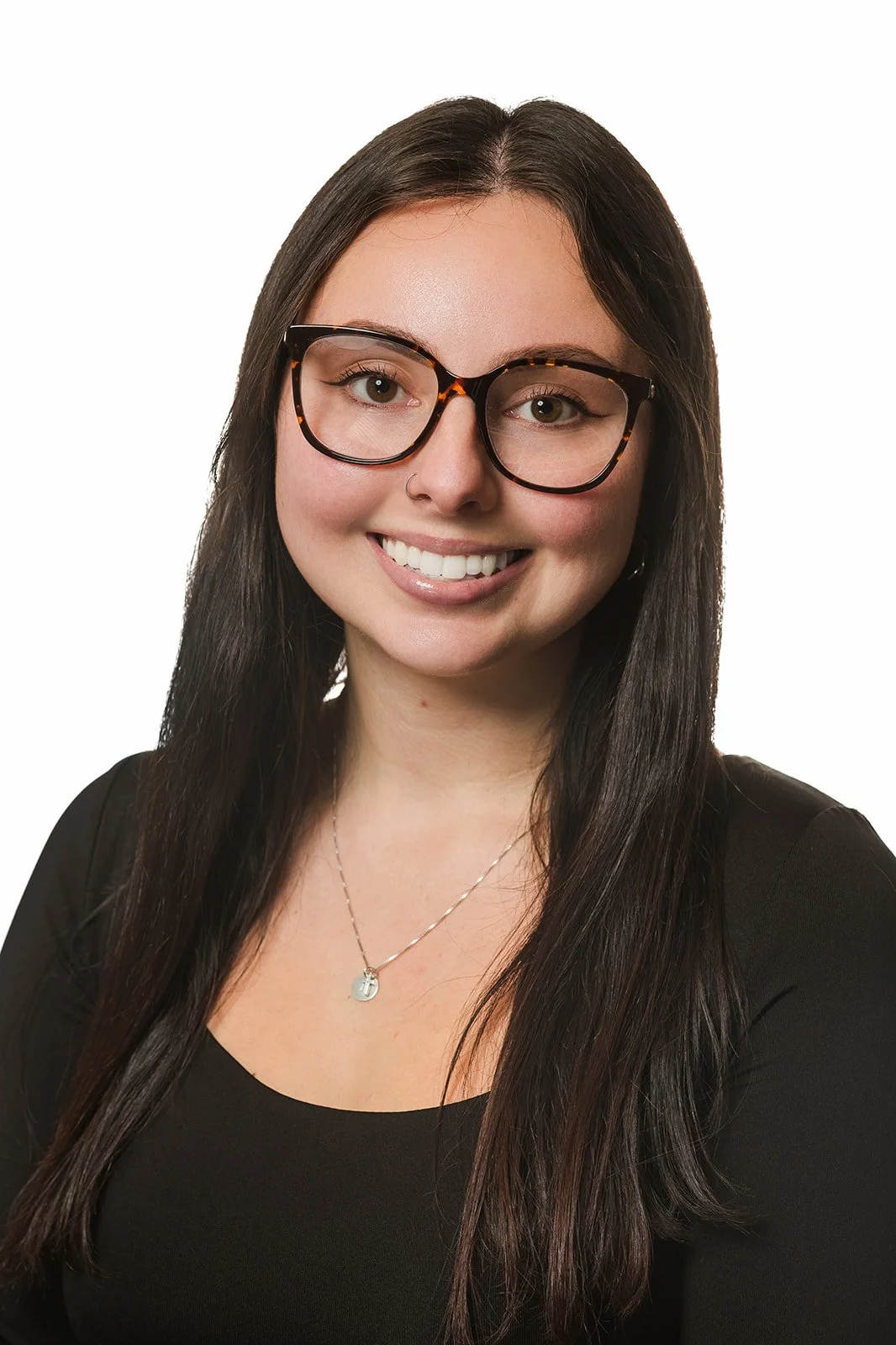 A young woman with long dark hair wearing glasses, a small nose ring, a black top, and a silver necklace, smiling against a white background.