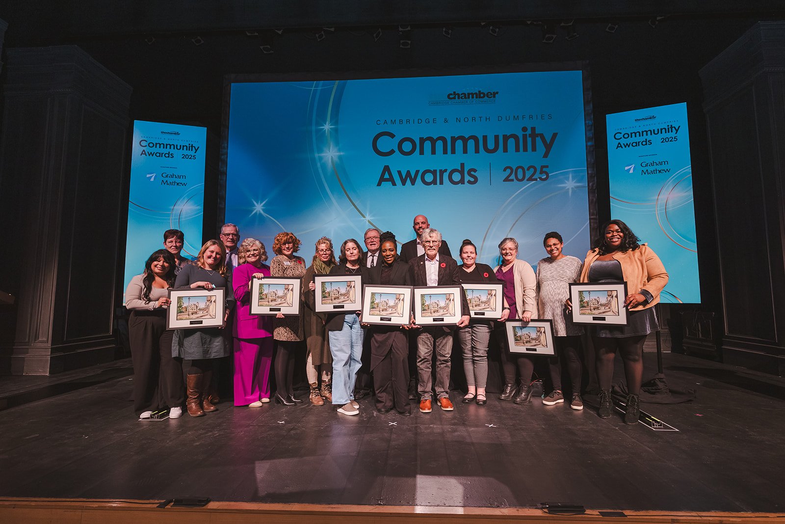 Group of people on stage holding framed awards at the 2025 Cambridge & North Dumfries Community Awards, with a large screen displaying the event details behind them.
