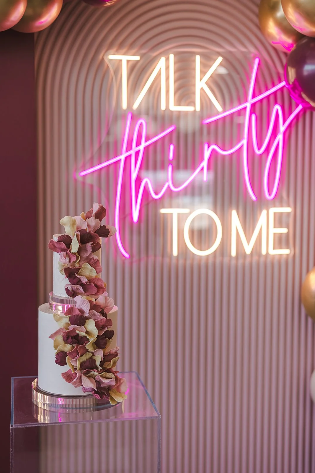 A white cake with pink, purple, and cream-colored petals cascading down the side is displayed on a clear stand. Behind the cake, there is a pink neon sign that reads 'TALK to him THANK YOU' in cursive and block letters, mounted on a background with vertical pink and beige stripes.