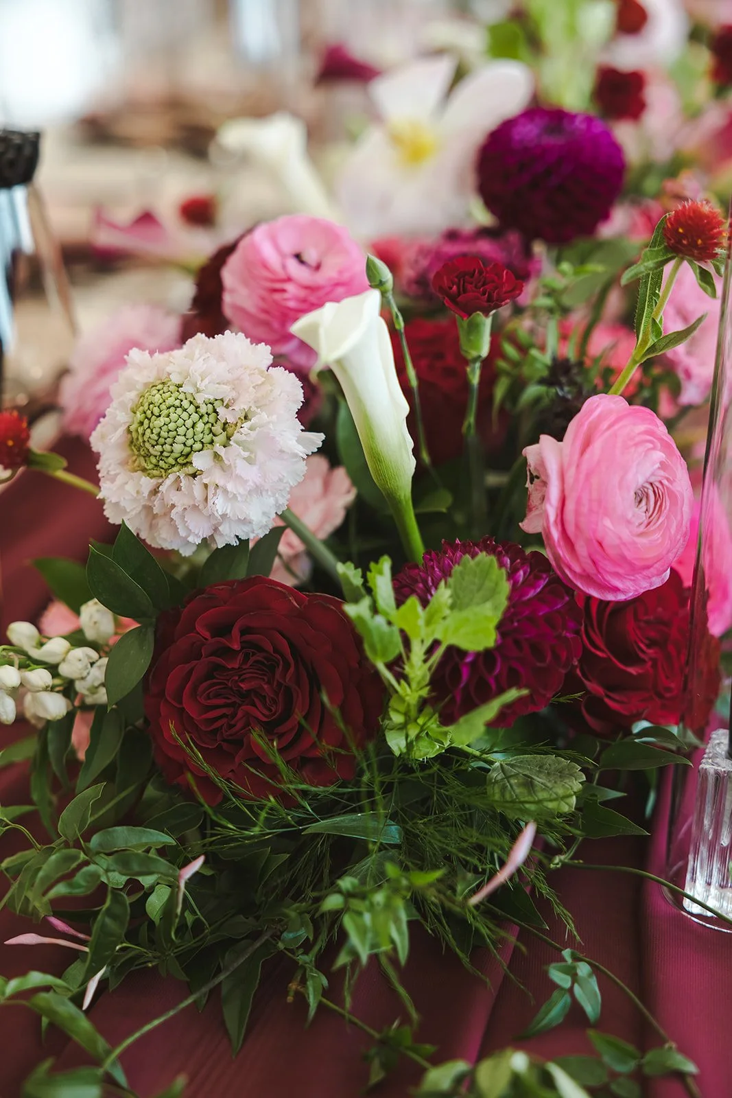 An arrangement of various flowers including pink ranunculus, white calla lily, and deep red roses with green foliage.