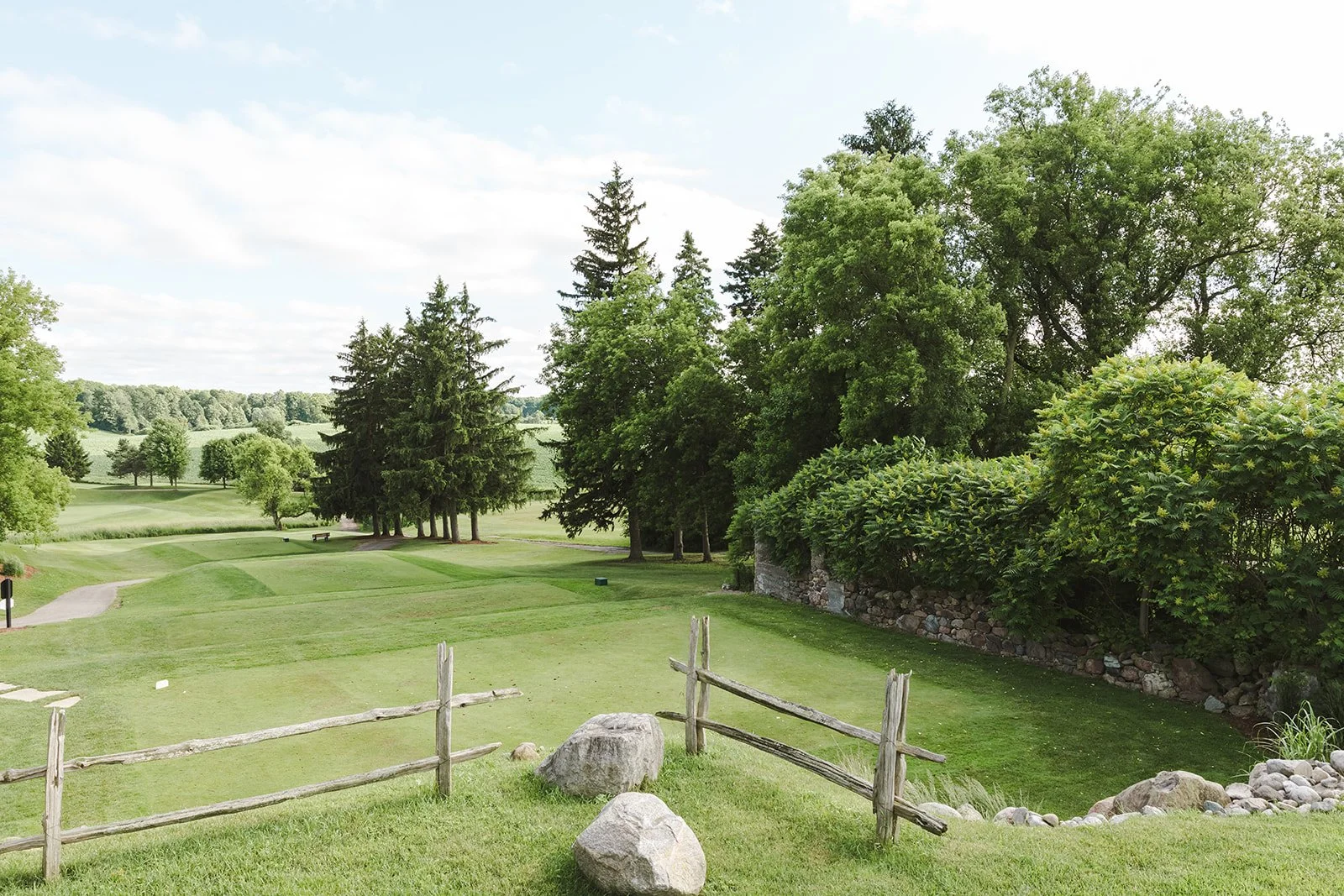 Scenic view of a golf course with lush green grass, trees, a stone wall, and a rustic wooden fence in a rural setting on a partly cloudy day.