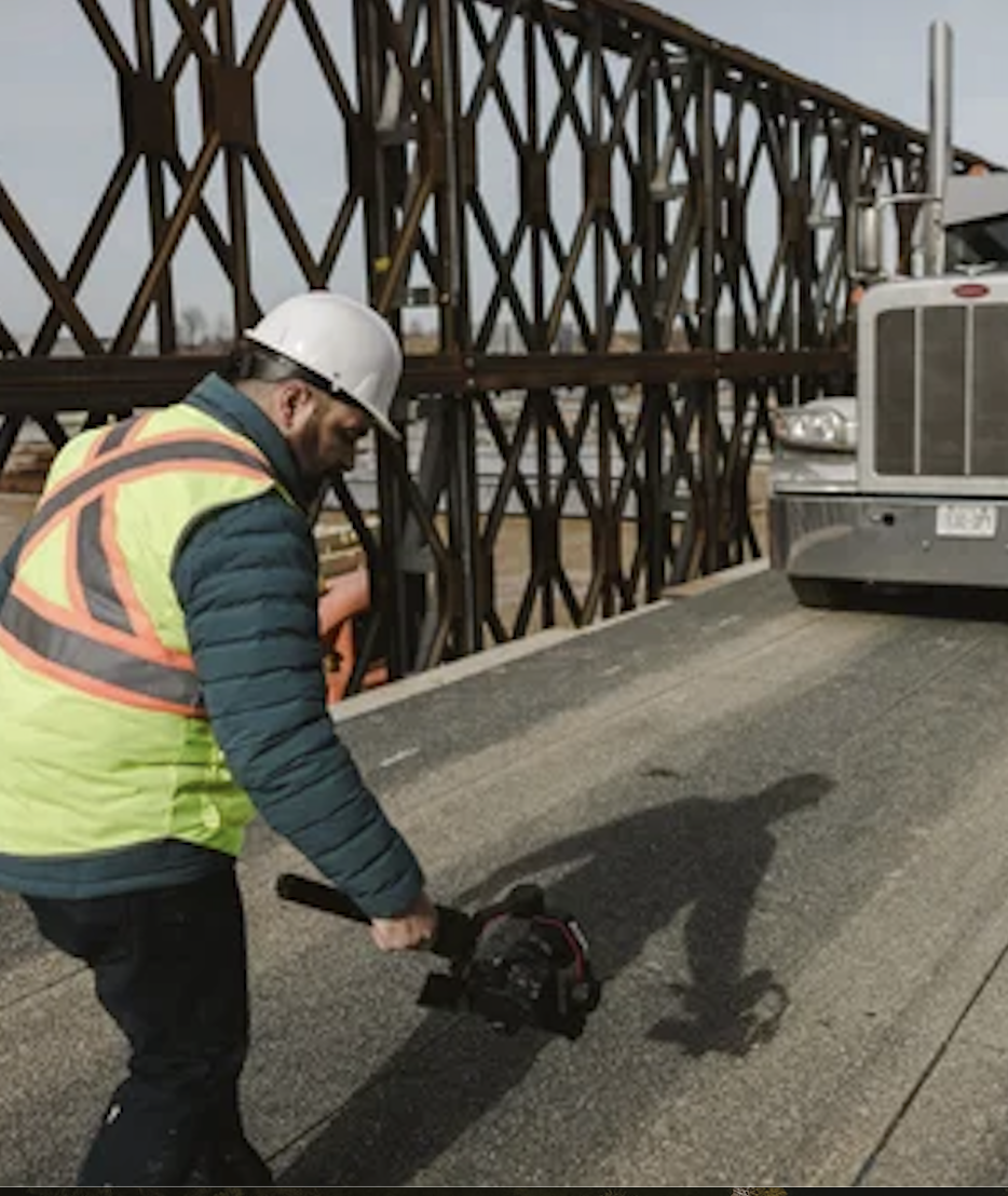 Construction worker using a handheld power saw on an asphalt surface, wearing a white hard hat and yellow safety vest, with a steel structure and a semi-truck in the background.
