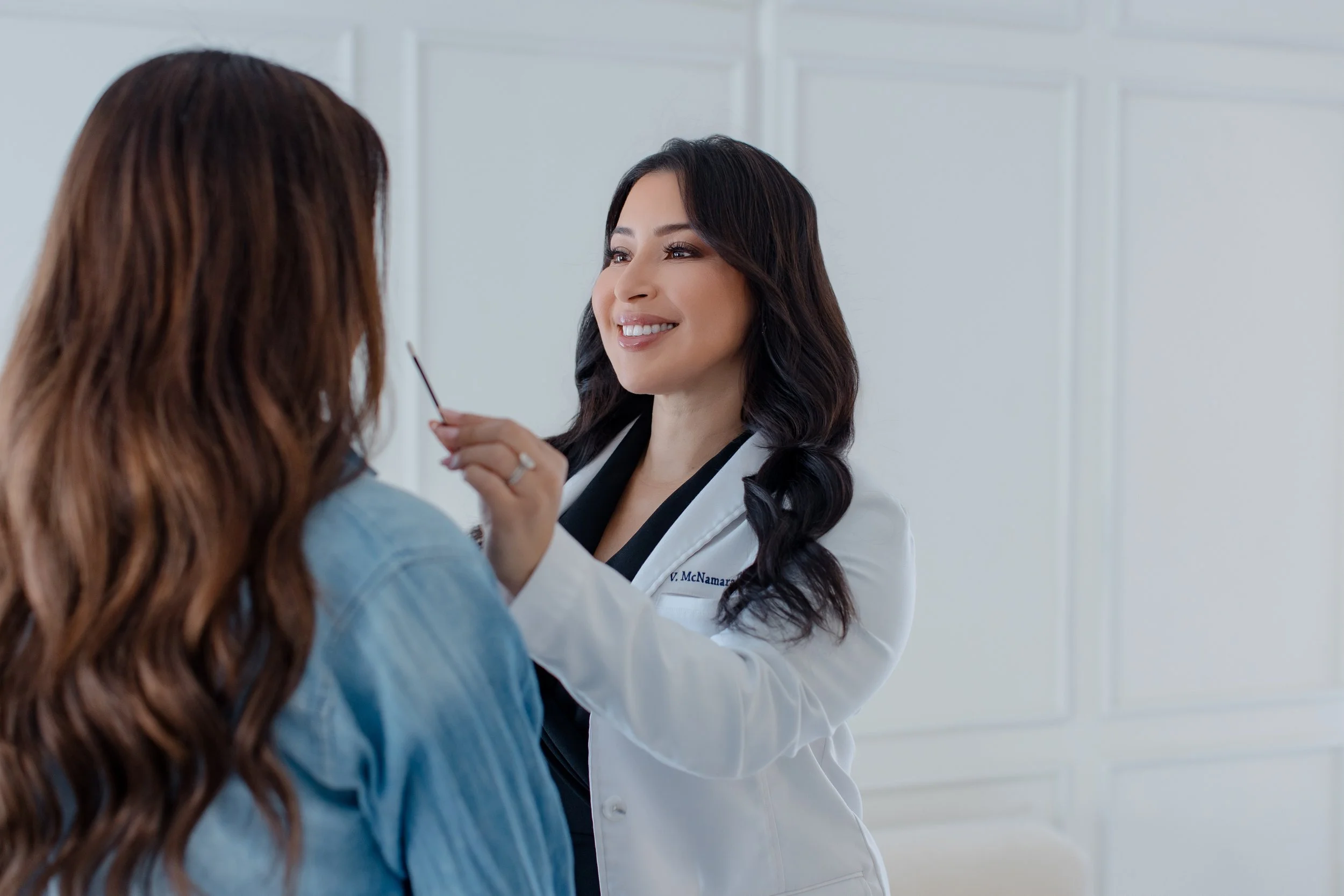 A female doctor with long dark hair smiling and holding a makeup brush near a patient with long reddish-brown hair. The patient is wearing a blue shirt. The doctor is dressed in a white coat.
