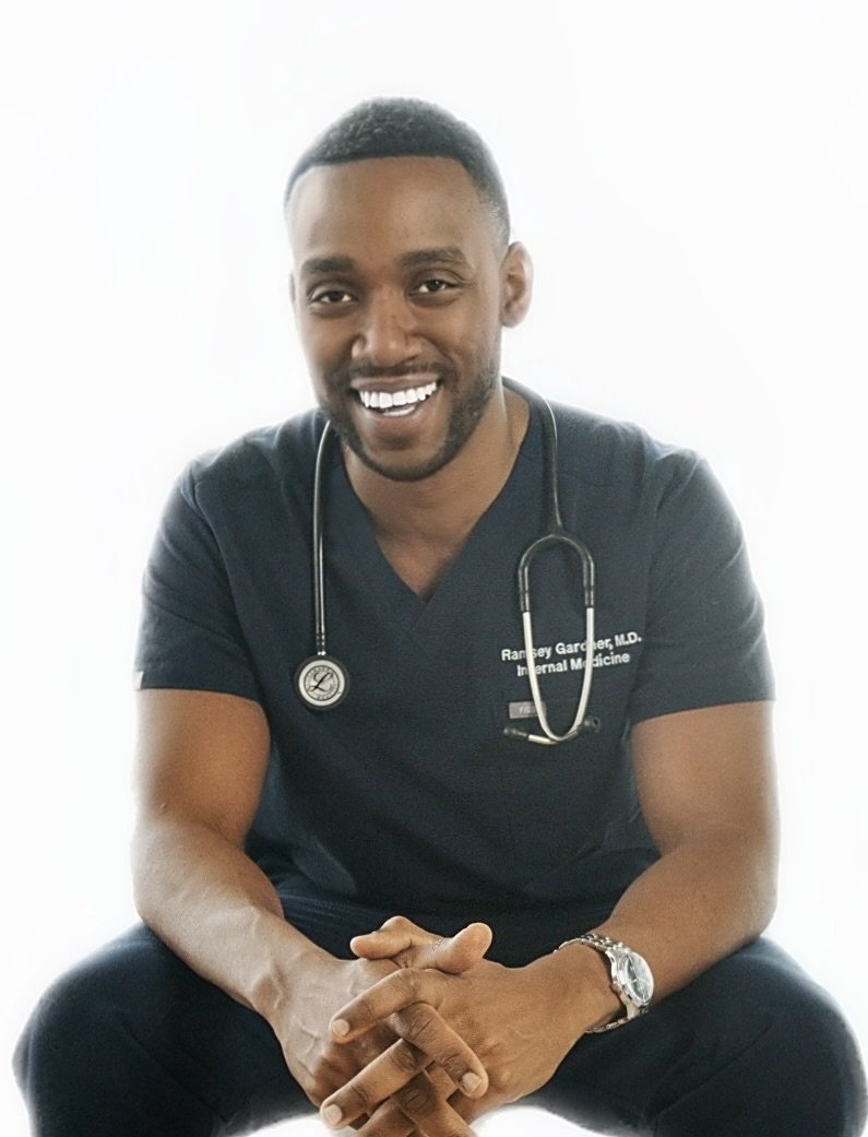 A smiling African American man wearing medical scrubs and a stethoscope around his neck sits with his hands clasped in front of him, against a plain white background.