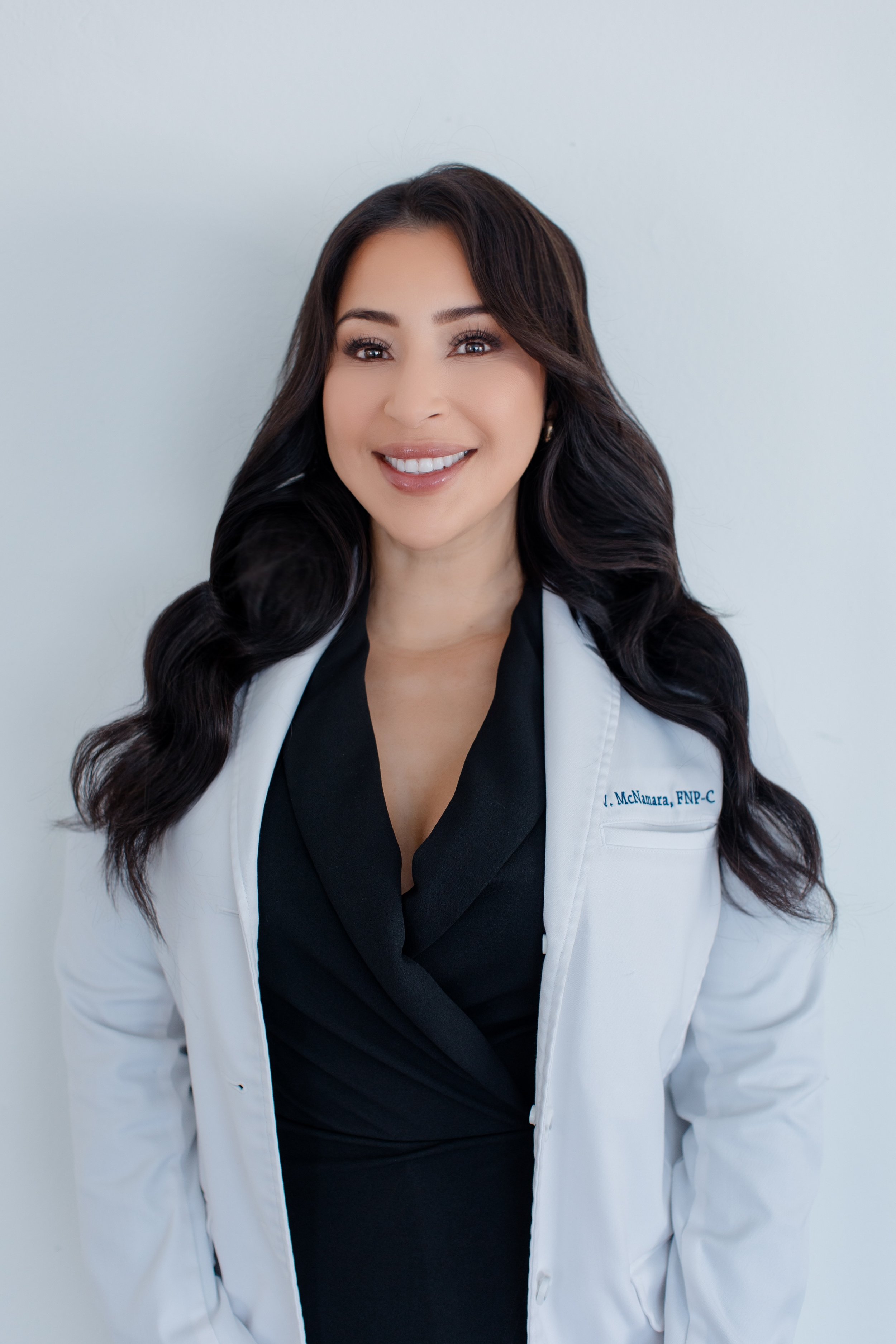 A woman with long dark wavy hair smiling, wearing a white coat with a name tag and a black top, standing against a plain light-colored background.
