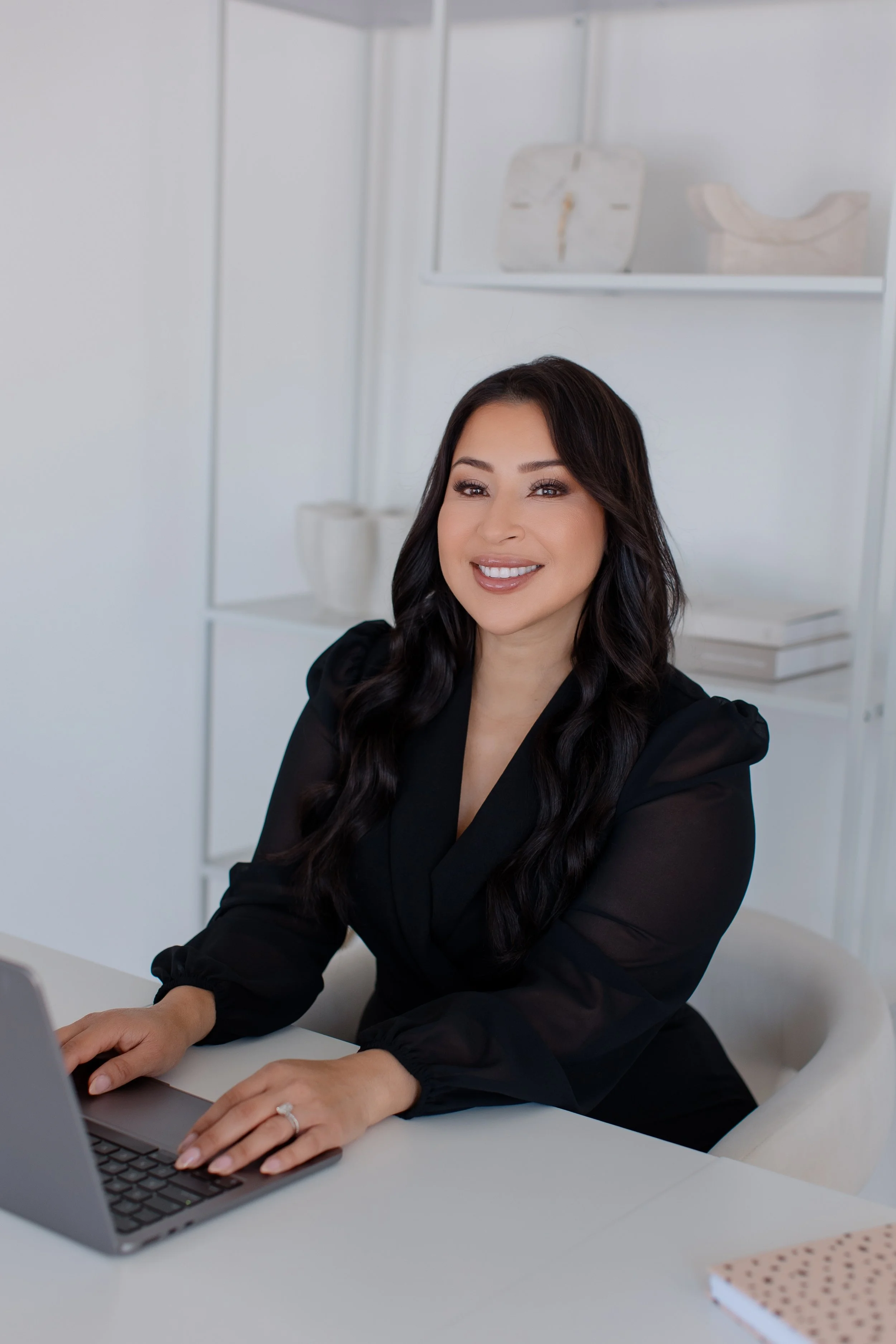 A woman sitting at a desk with a laptop, smiling, in a modern office space with white shelves and decorative items in the background.