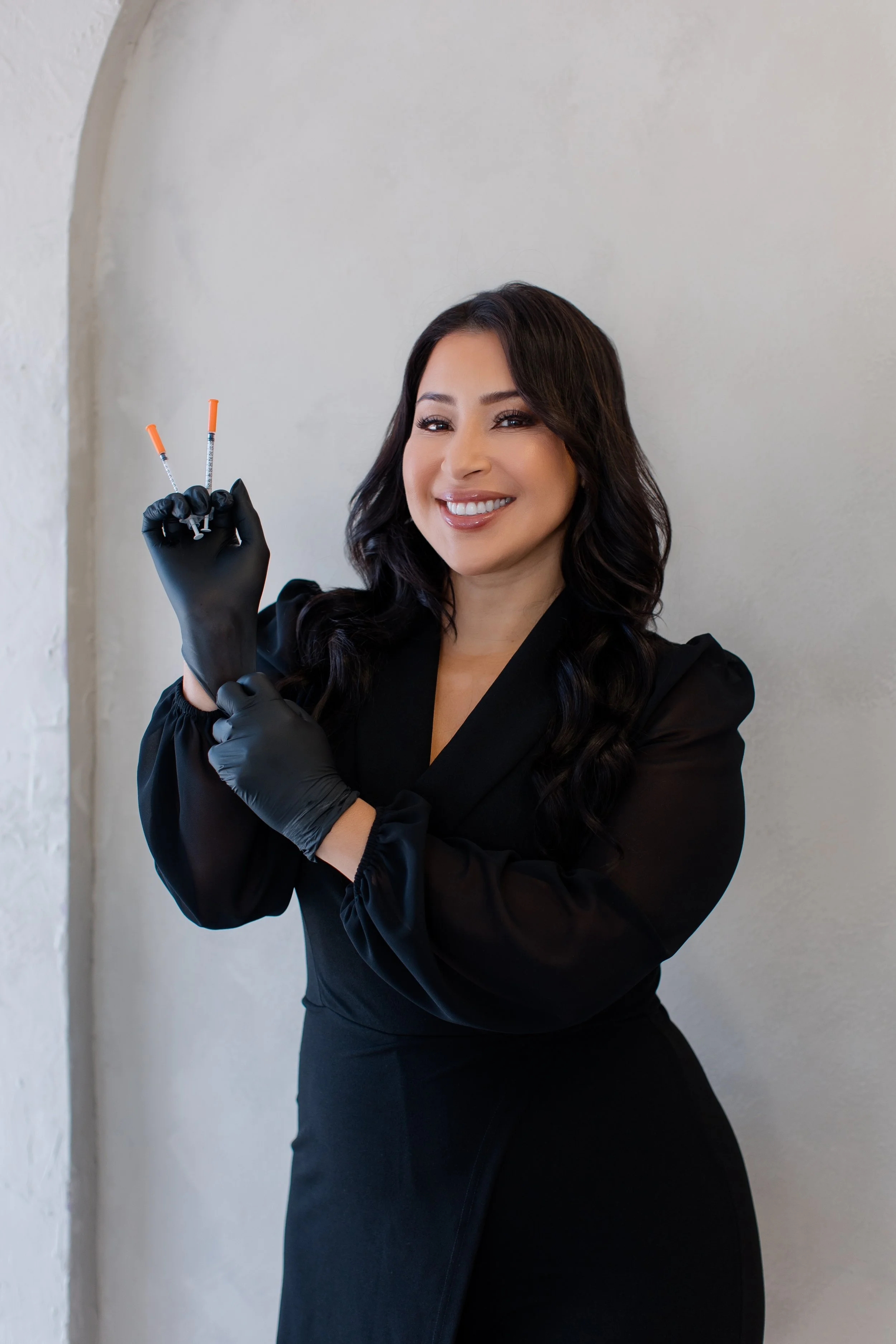 A woman with dark wavy hair smiling, wearing black gloves and a black dress, holding two syringes.