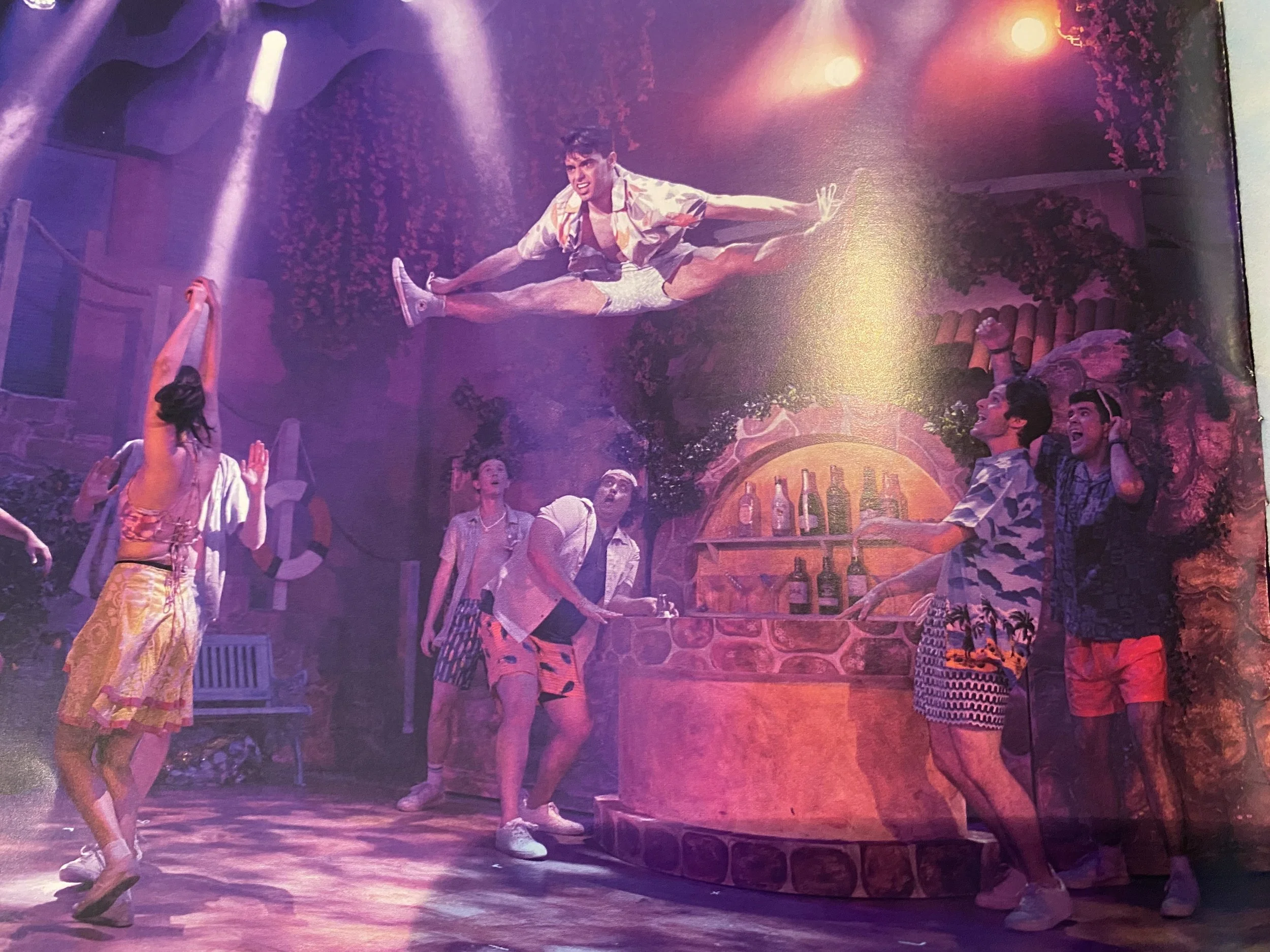Actors performing a dance routine from Mamma Mia in 2025 surrounded by colorful set decorations and ensemble performers.