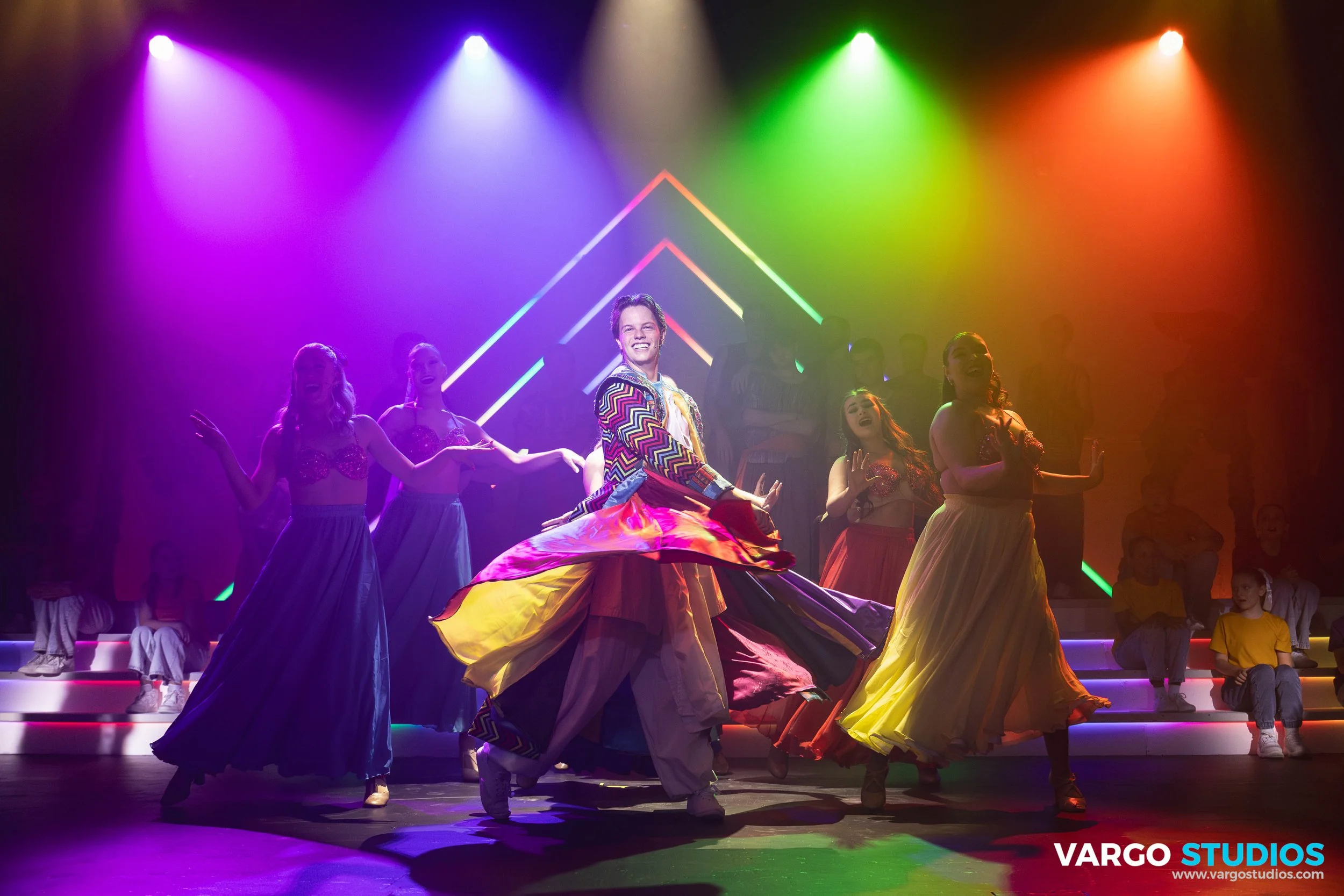 Stage performance with dancers in colorful flowing skirts and a male performer in a patterned jacket, under vibrant rainbow lights.