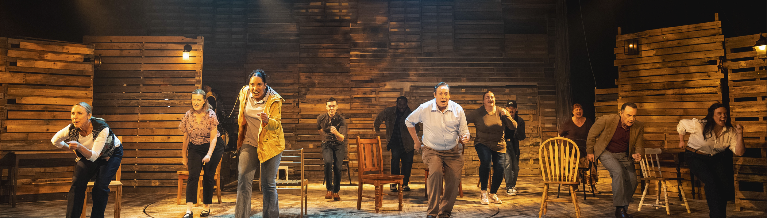 A group of people performing on a stage with wooden background, some standing and some sitting, engaged in an energetic activity or acting.