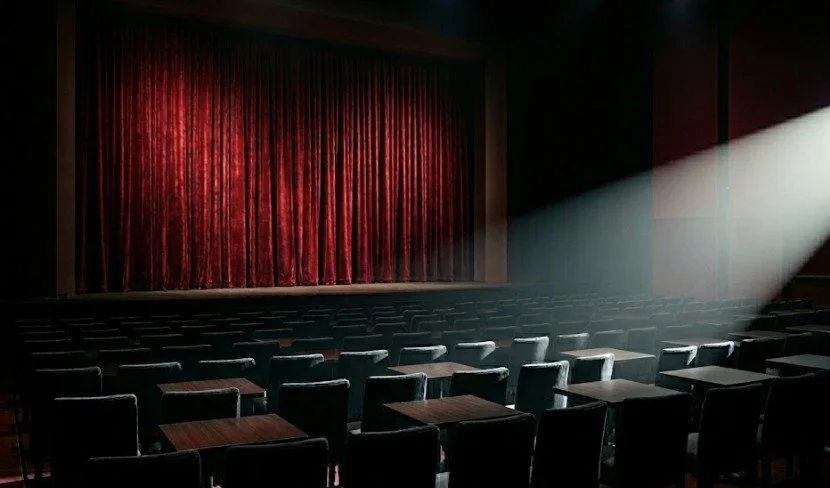 Empty theater with closed red velvet curtains and a single spotlight illuminating the seats.