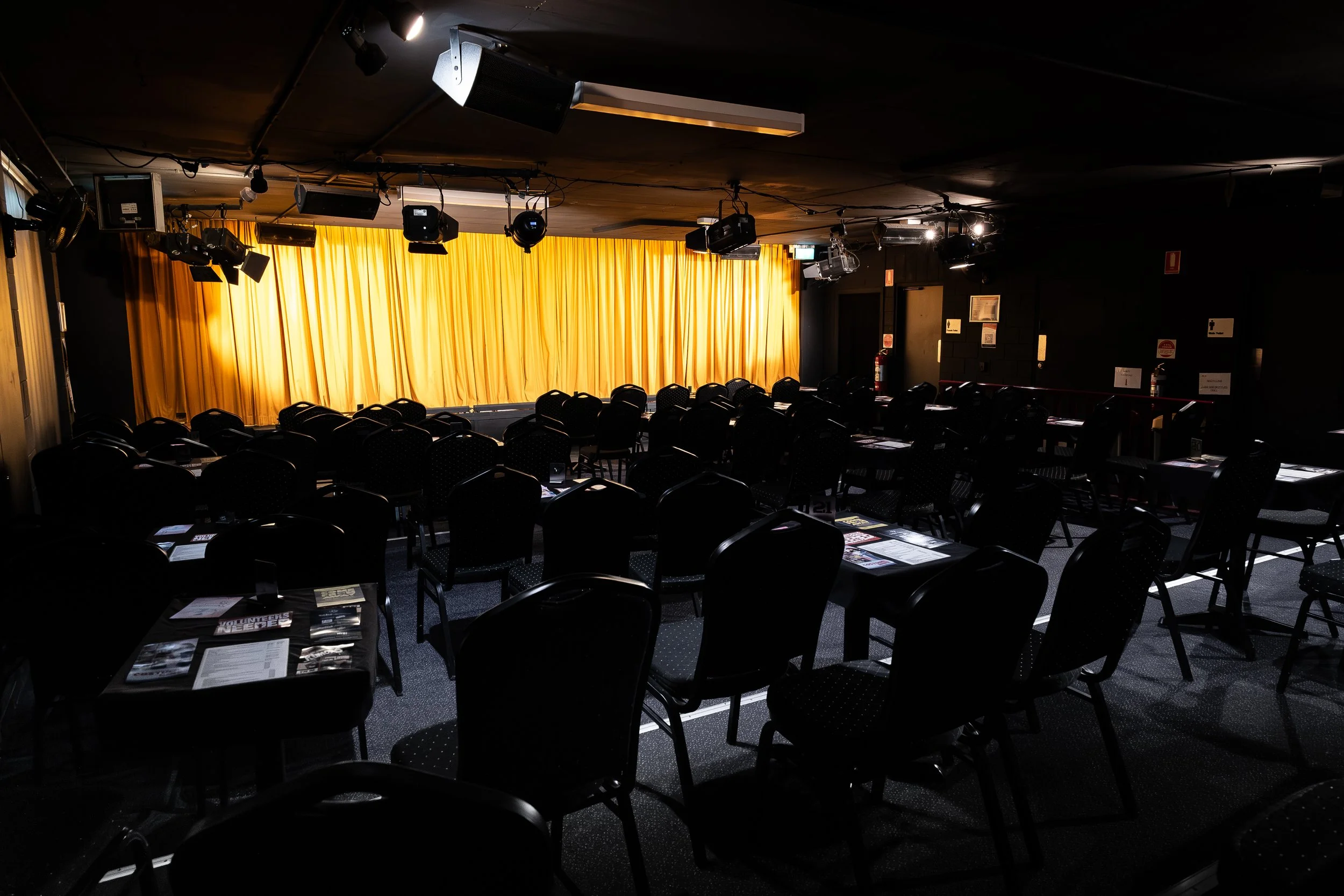 Empty theater or auditorium with rows of chairs, a stage with a yellow curtain, and various stage lights and equipment hanging from the ceiling.