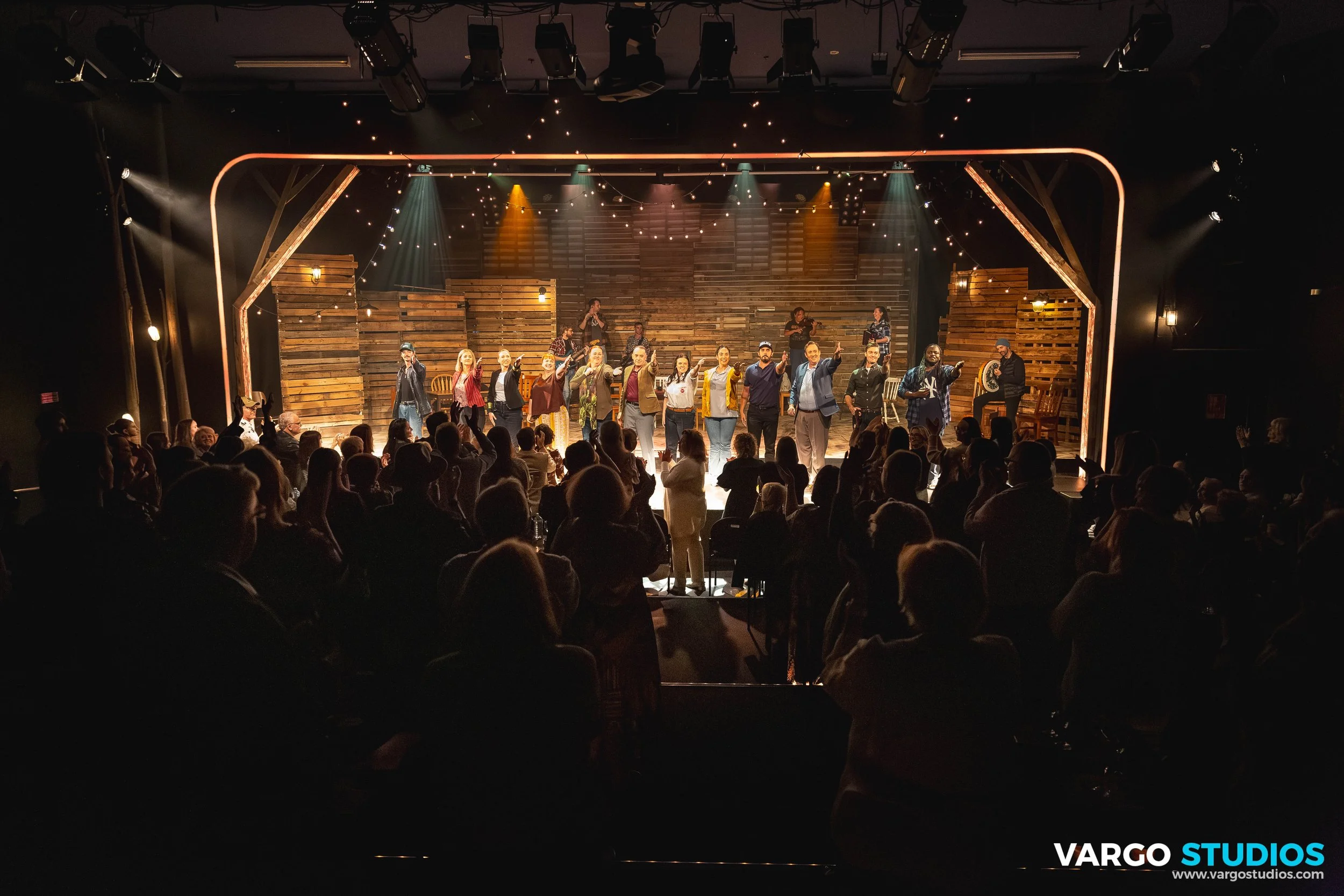 Stage performance at a theater with a group of actors holding hands, audience members clapping, stage decorated with wooden pallets and string lights, with lighting fixtures overhead.