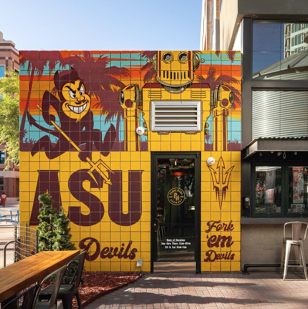 Colorful mural on a brick building featuring a mischievous cartoon devil holding pitchfork, a retro robot head, and palm trees in the background. The mural has large text reading "ASU" and "Devils," indicating it's for Arizona State University. The building has a door with a sign displaying hours and an adjacent window.