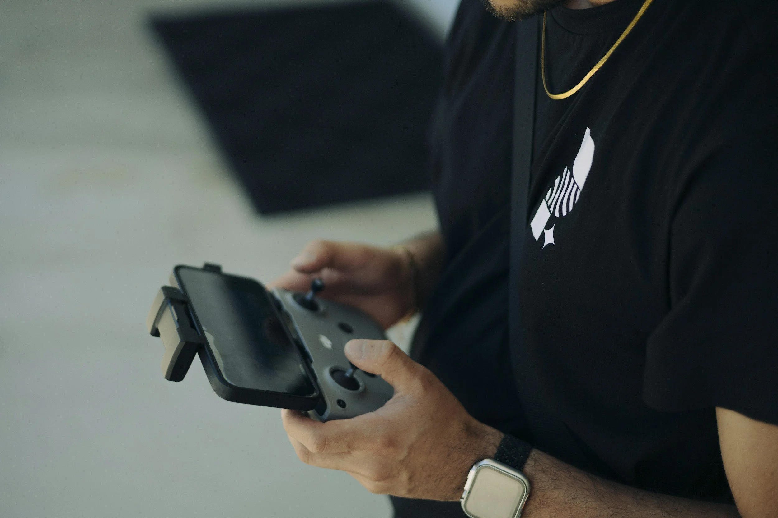 Person holding a drone remote controller, wearing a black shirt with a logo, a gold chain, and a smartwatch.