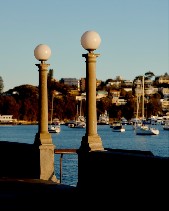 Two decorative lamps on stone pillars overlooking a marina with sailboats and a hillside with houses.