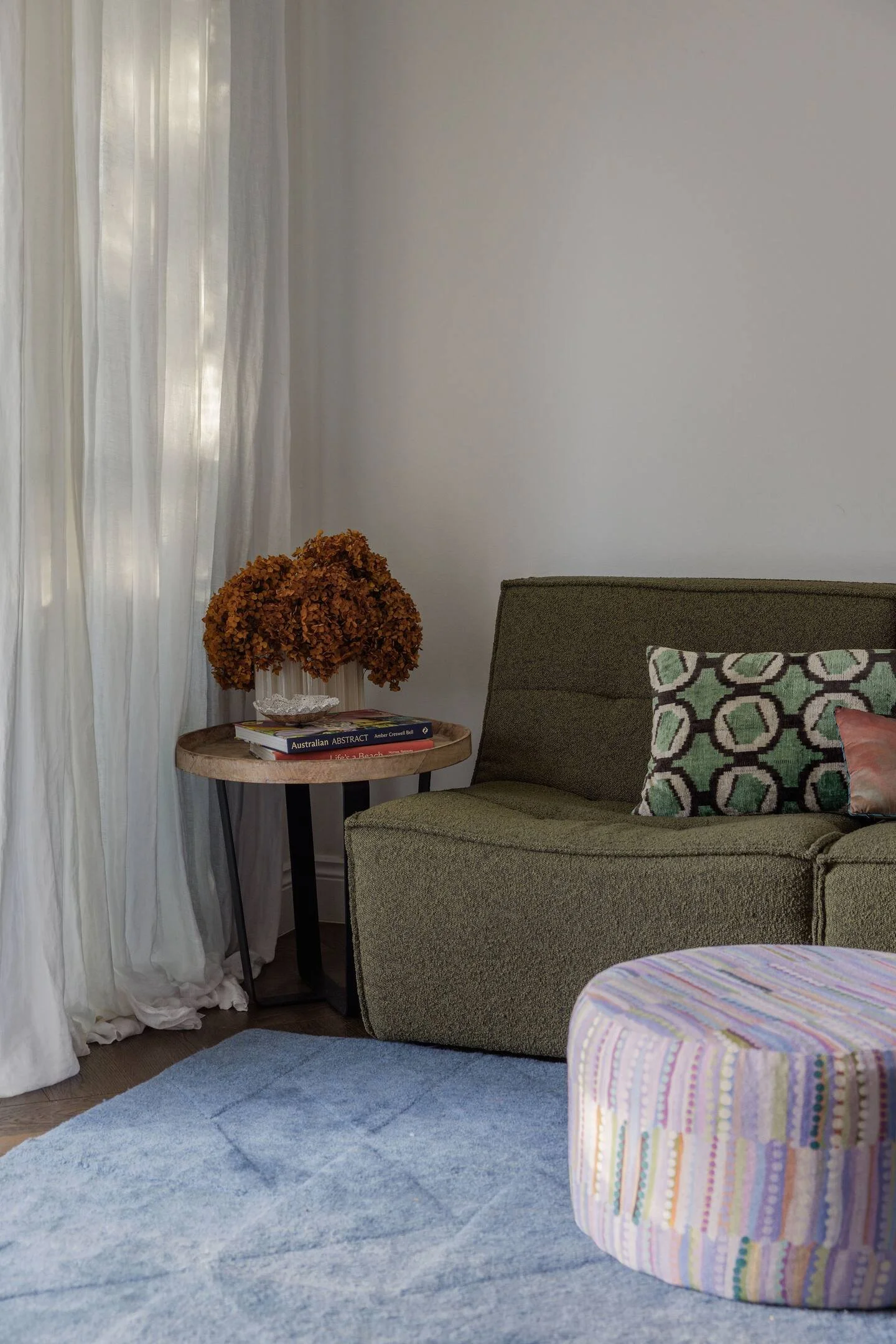 A cozy living room corner with a green sofa, a round side table holding books and a dried floral arrangement, white curtains, a light blue rug, and a colorful pouf.