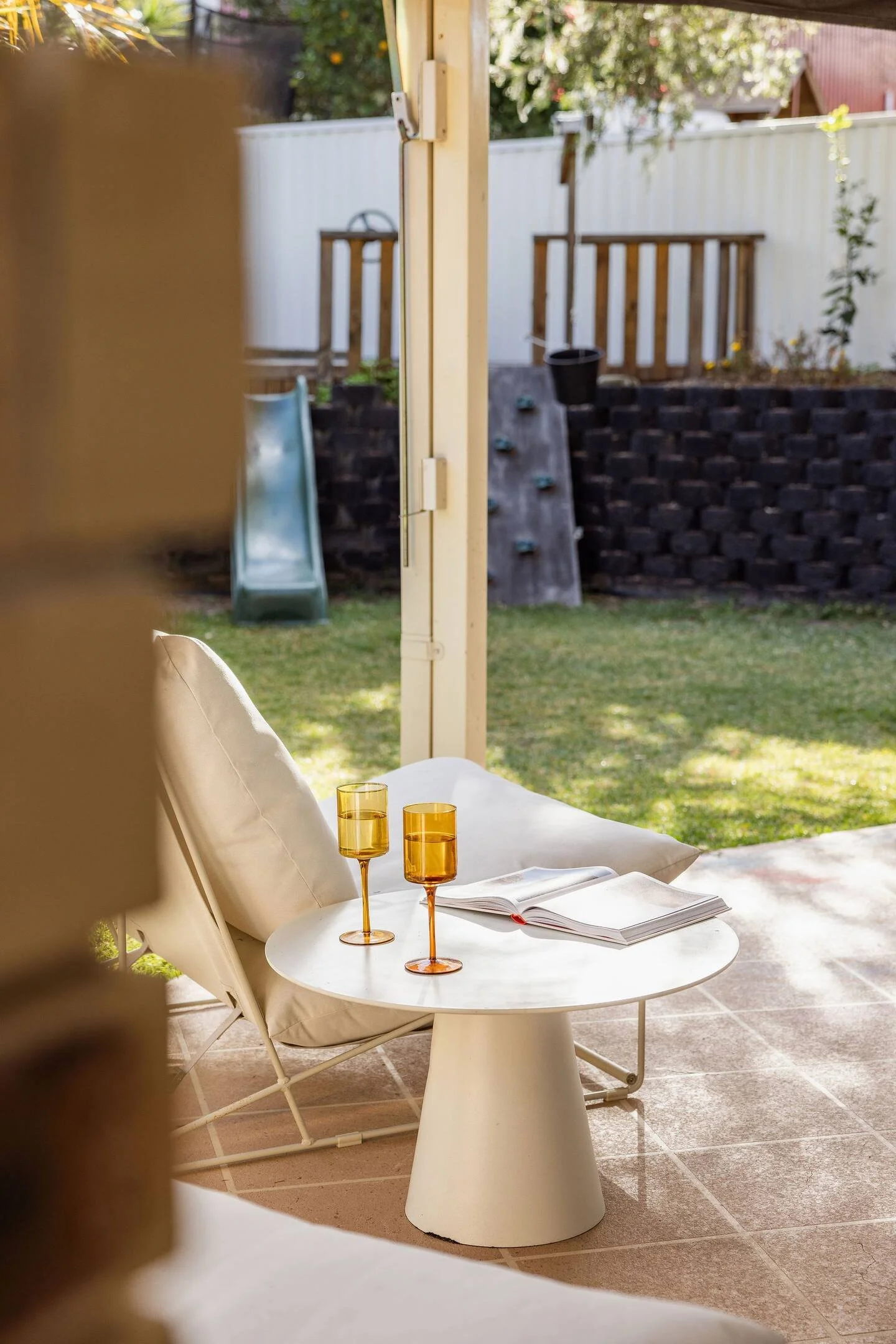 Outdoor patio setup with a white side table holding two amber glasses and an open book, under a canopy. In the background, a grassy yard with a slide, climbing wall, and fenced perimeter.