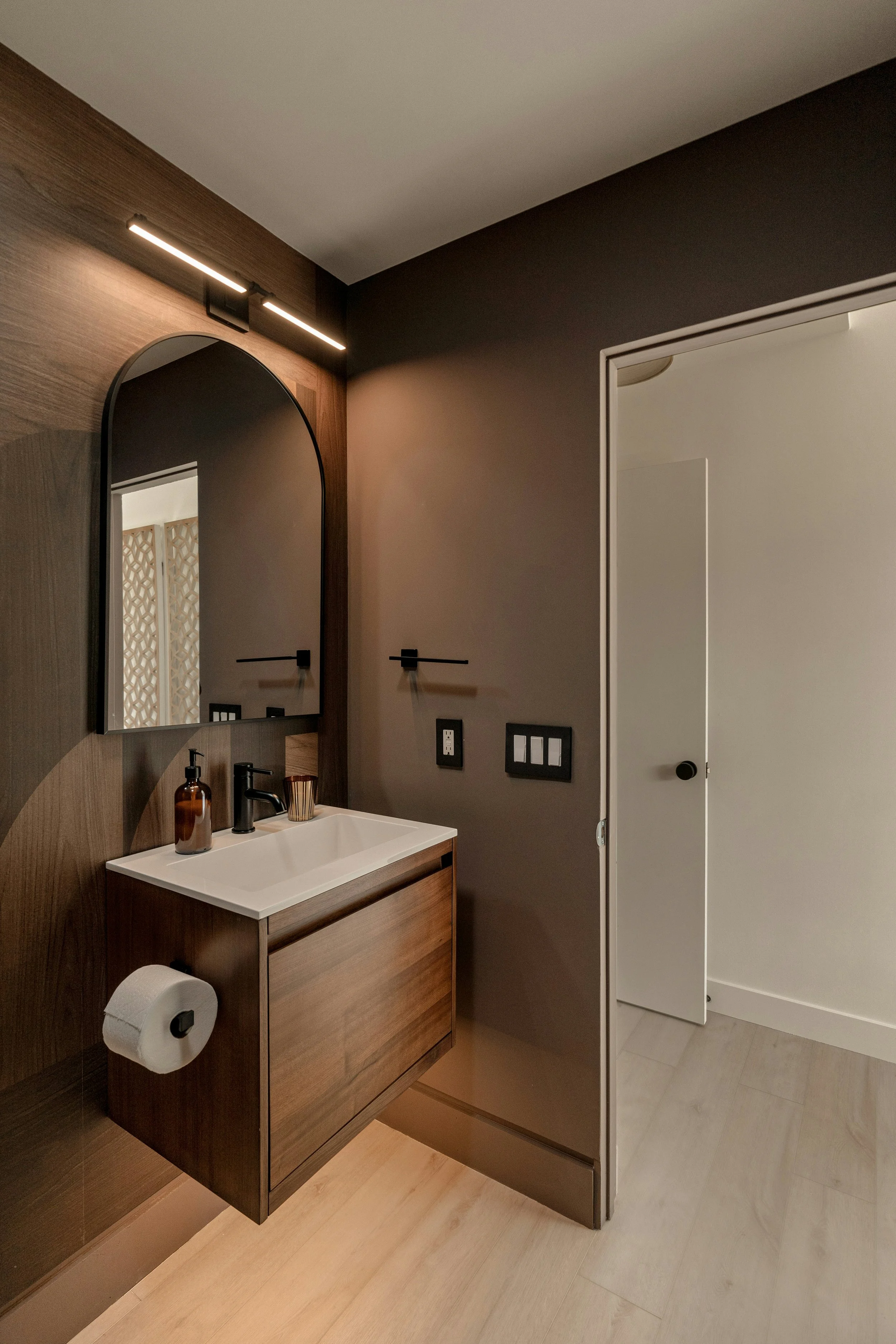Modern bathroom with a floating wooden vanity, a white sink, a black faucet, a large arched mirror, and a light fixture overhead. There is a wall-mounted toilet paper holder and a partially open door leading to another room.