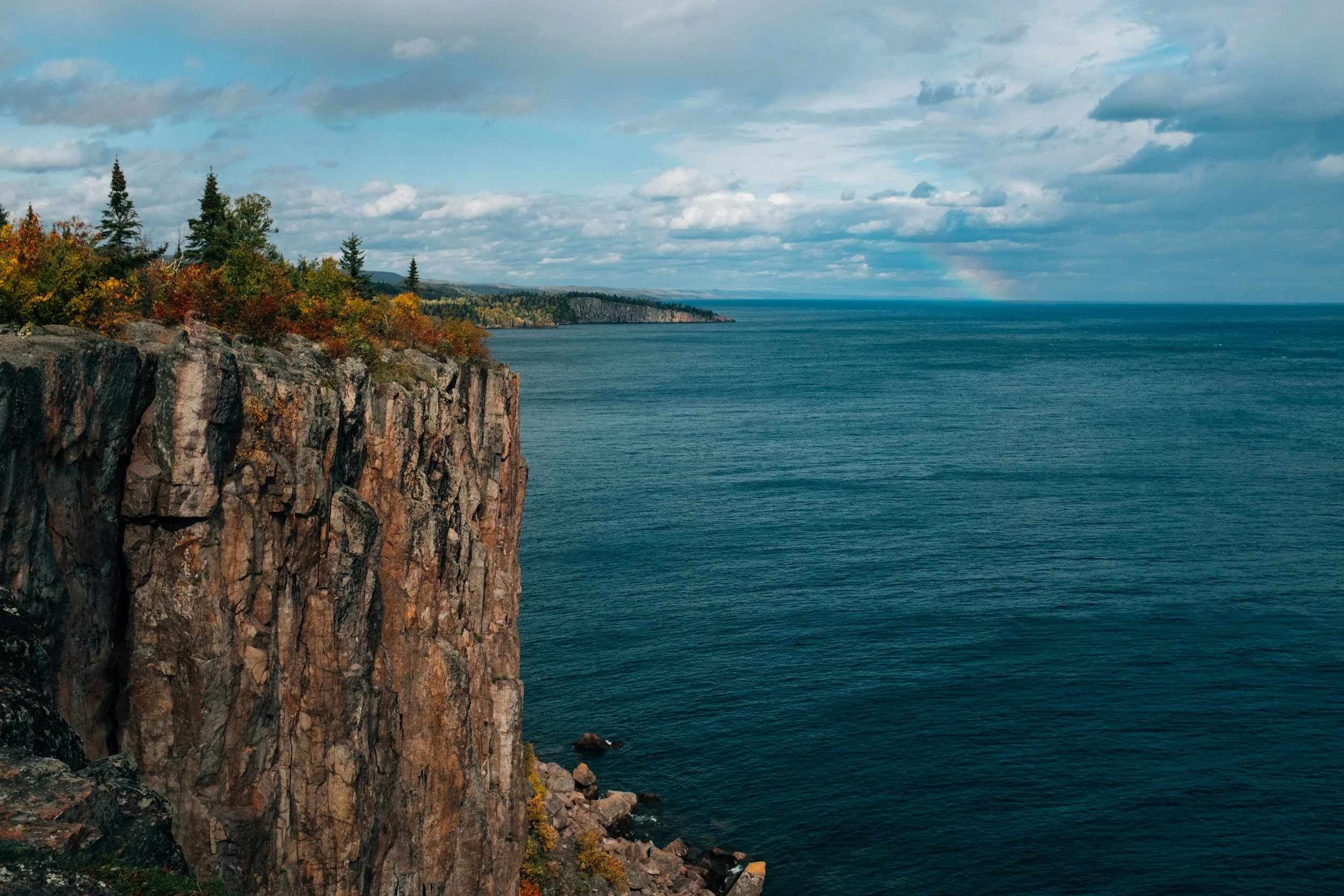 Cliffside with rocky edge, trees, and colorful fall foliage overlooking a large body of water with a cloudy sky and a faint rainbow in the distance.