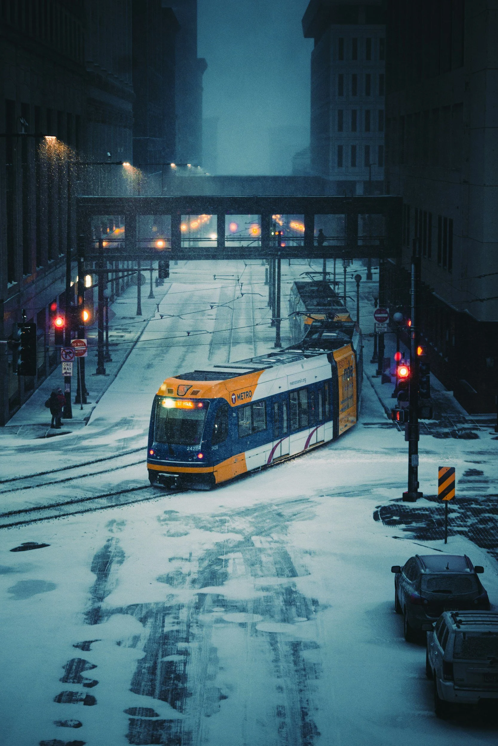 A yellow and blue metro tram driving on snow-covered city street at night, with traffic lights and tall buildings in the background.