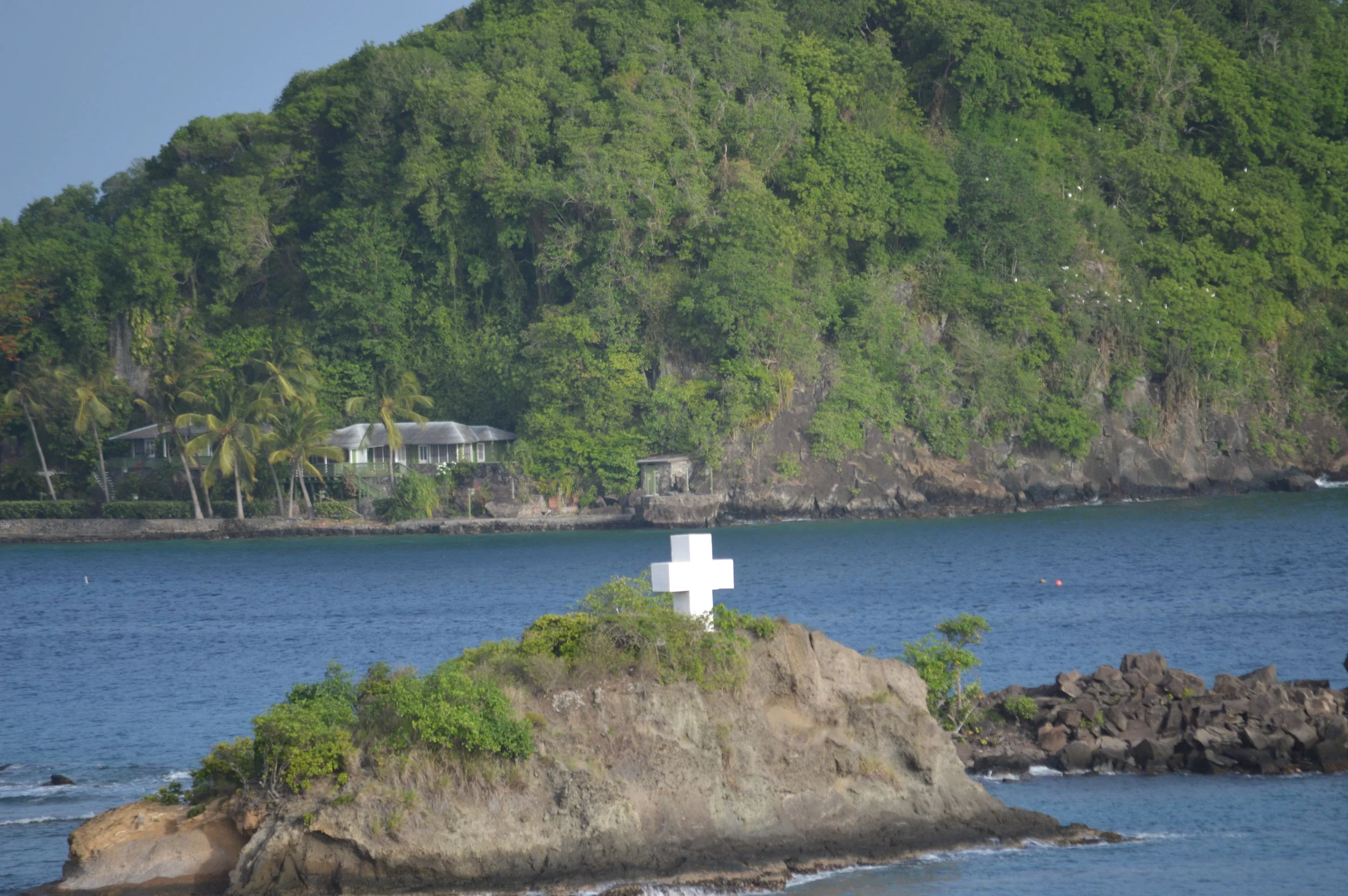 A white cross on a small rocky island surrounded by water, with green bushes and trees, and a hillside covered with lush green foliage and a building with a white roof in the background.