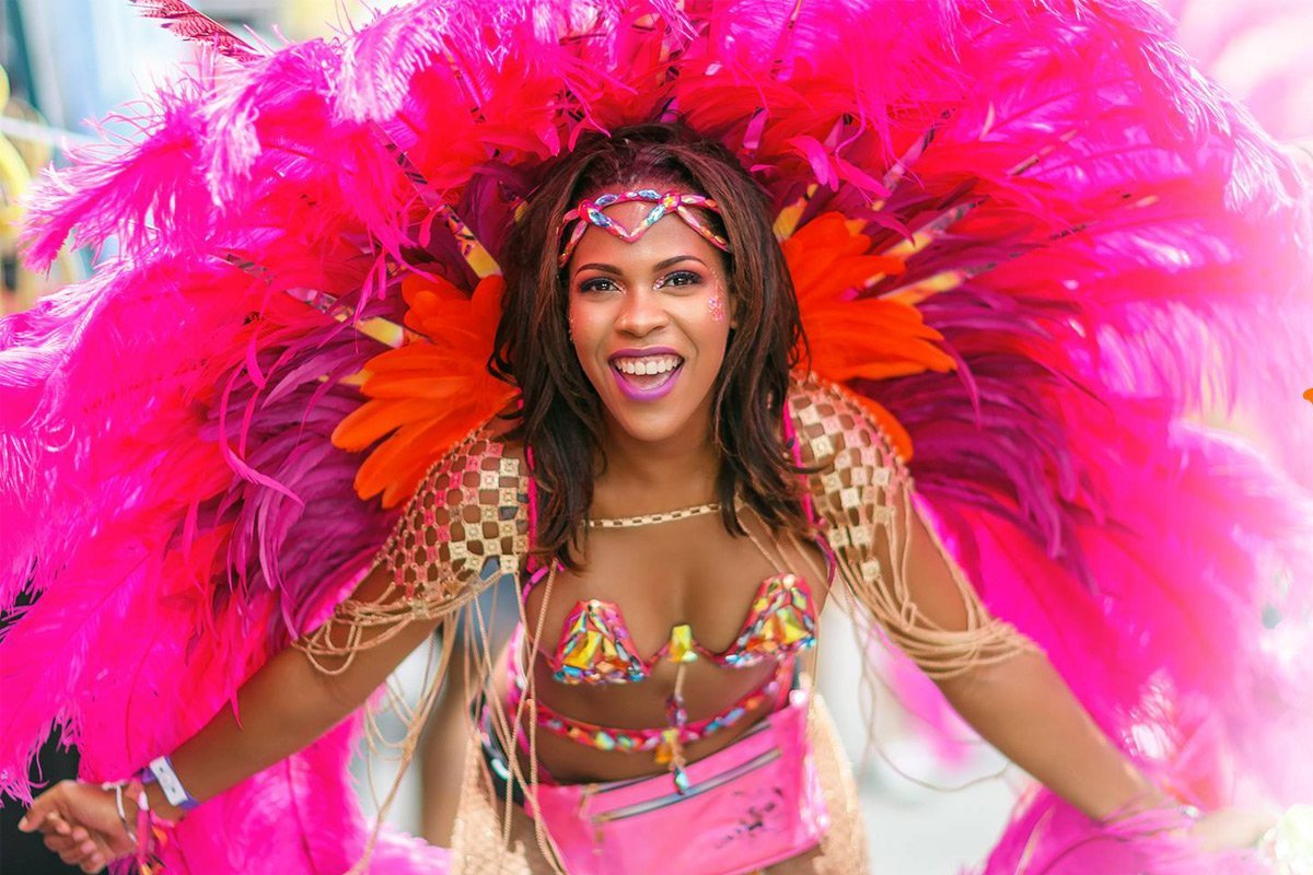 A woman in a colorful carnival costume with pink feathers, orange accents, and a beaded bikini top, smiling and having fun.