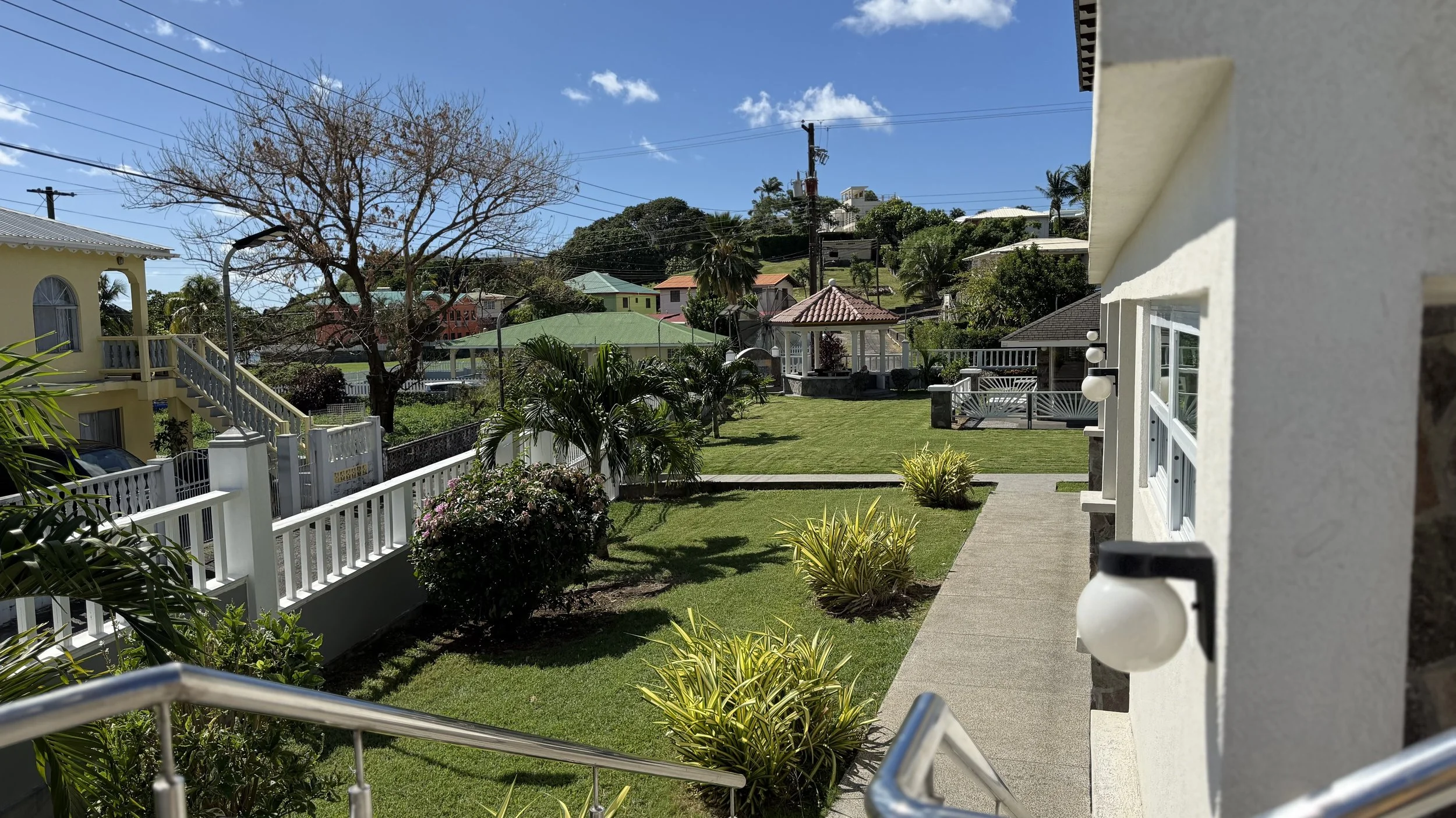 View of a landscaped backyard with a pathway, green grass, various trees and plants, a gazebo towards the center-back, and colorful houses on a hill in the background under a blue sky with some clouds.
