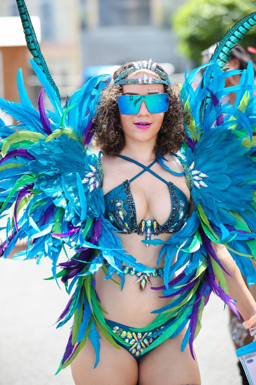 Woman in colorful carnival costume with large blue, green, purple feathers, wearing sunglasses, and a decorated bikini top and bottom.