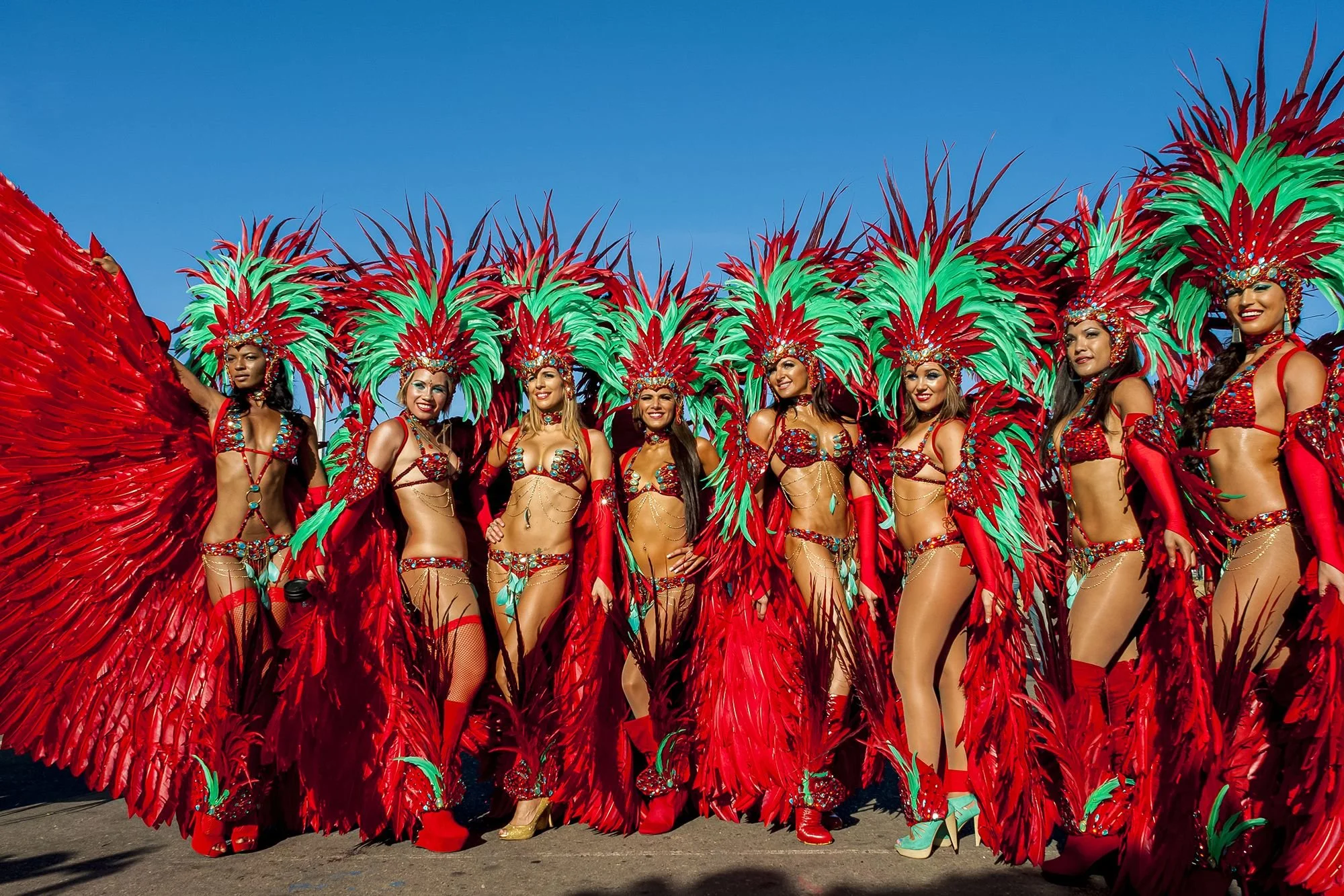 Group of women in colorful parade costumes with large feathered headdresses and matching outfits, standing outdoors against a clear blue sky.