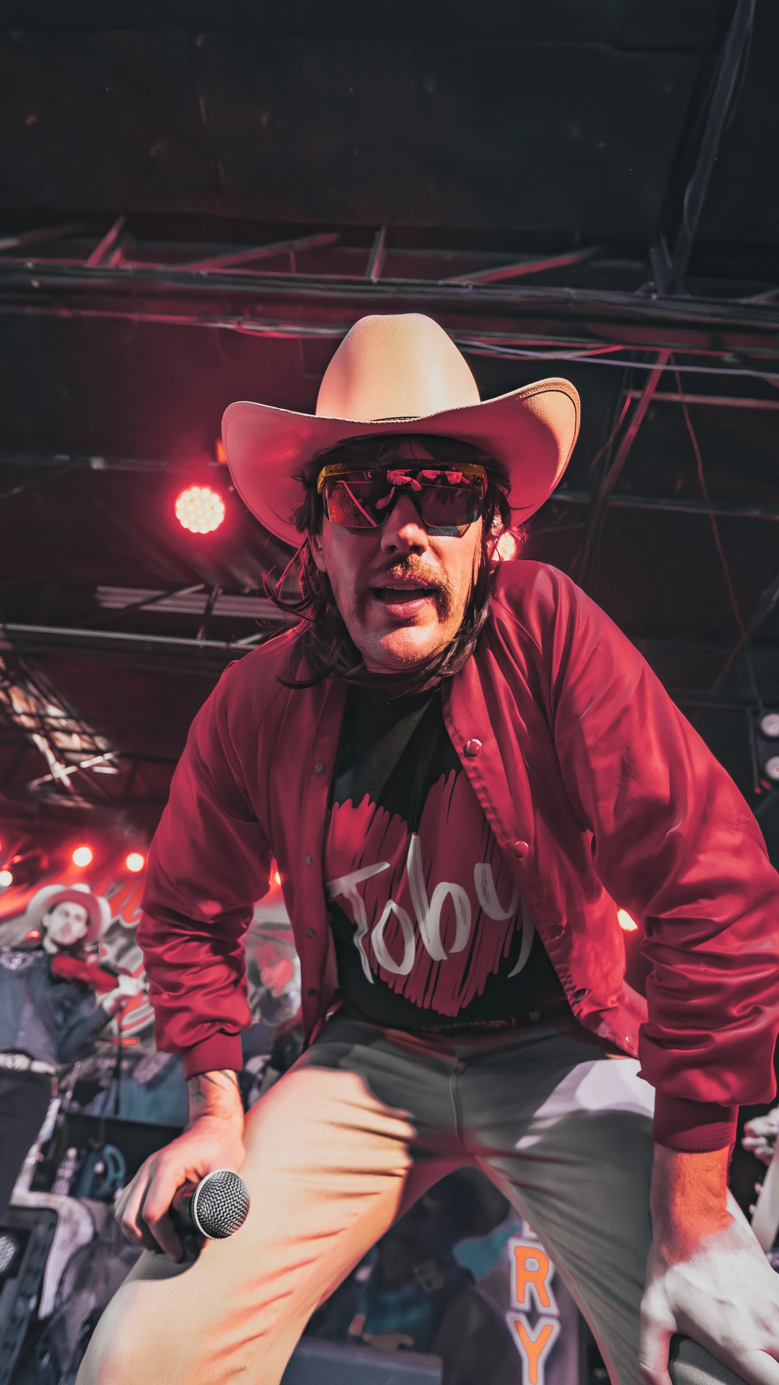 A man dressed in a cowboy hat, sunglasses, and a red jacket performing on stage with a microphone, with animated background and stage lighting.