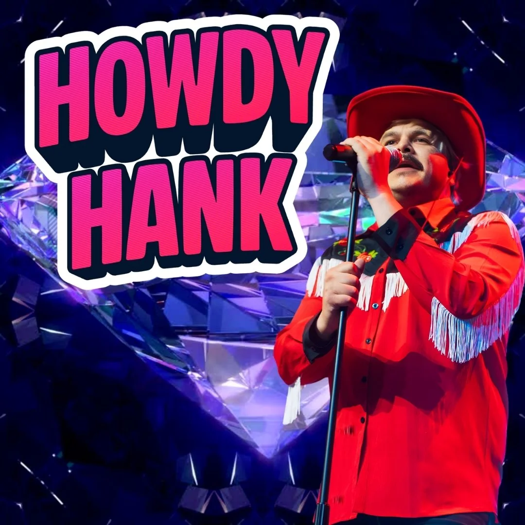 A man dressed in a red western shirt with white fringe details, wearing a matching red cowboy hat, performs on stage holding a microphone. The stage background features a large, faceted purple gemstone and the words "Howdy Hank" in bold pink and black letters.