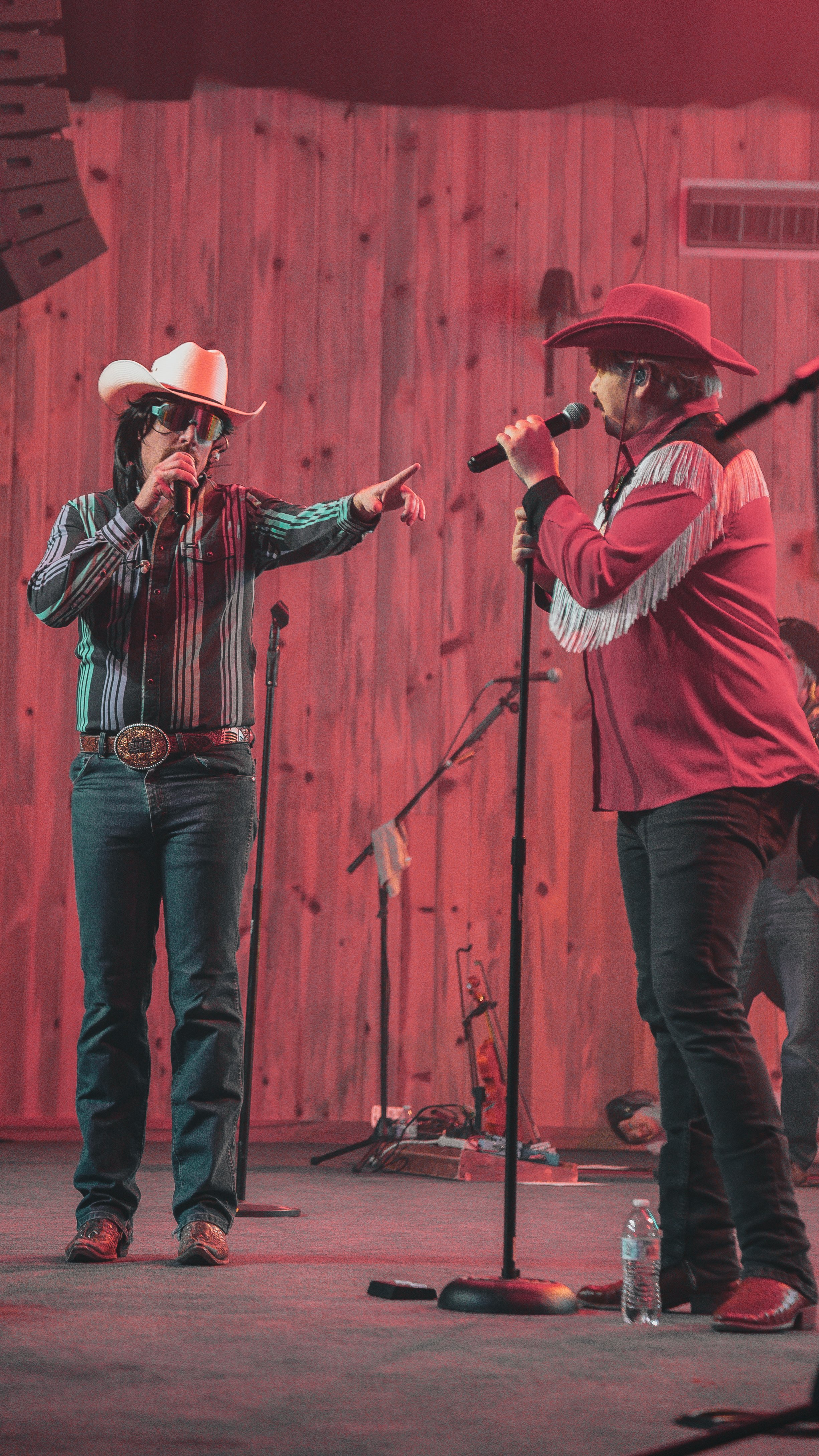 Two individuals dressed as country singers on stage, wearing cowboy hats and microphones, against a wooden backdrop, with stage equipment and a water bottle visible.