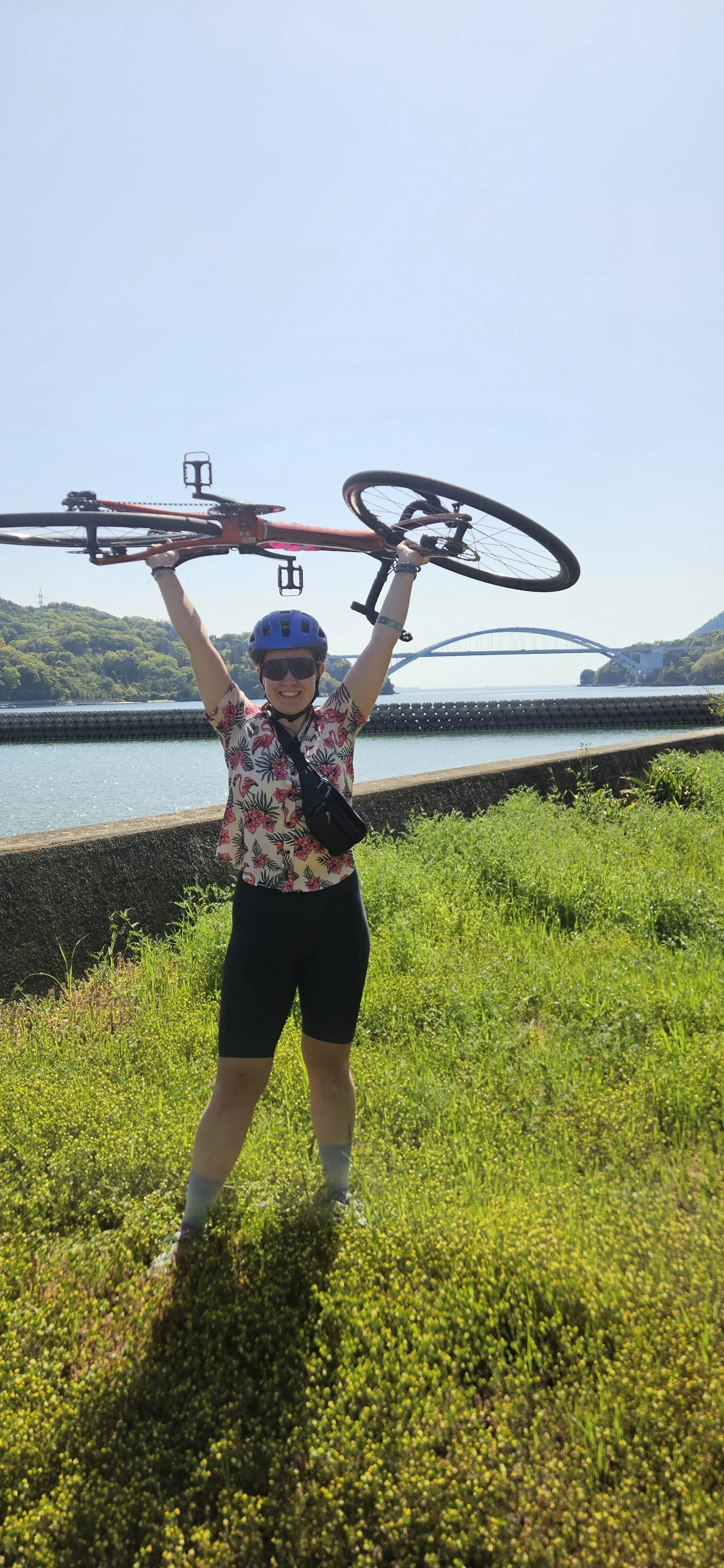 Chelle holds a road bike above her. She wears a button up shirt with flamingos, blue cycling helmet, black/purple sunglasses and black bib shorts. backdrop of a bridge along the Shimanami Kaido in Japan. Image by Gareth Brouwer.