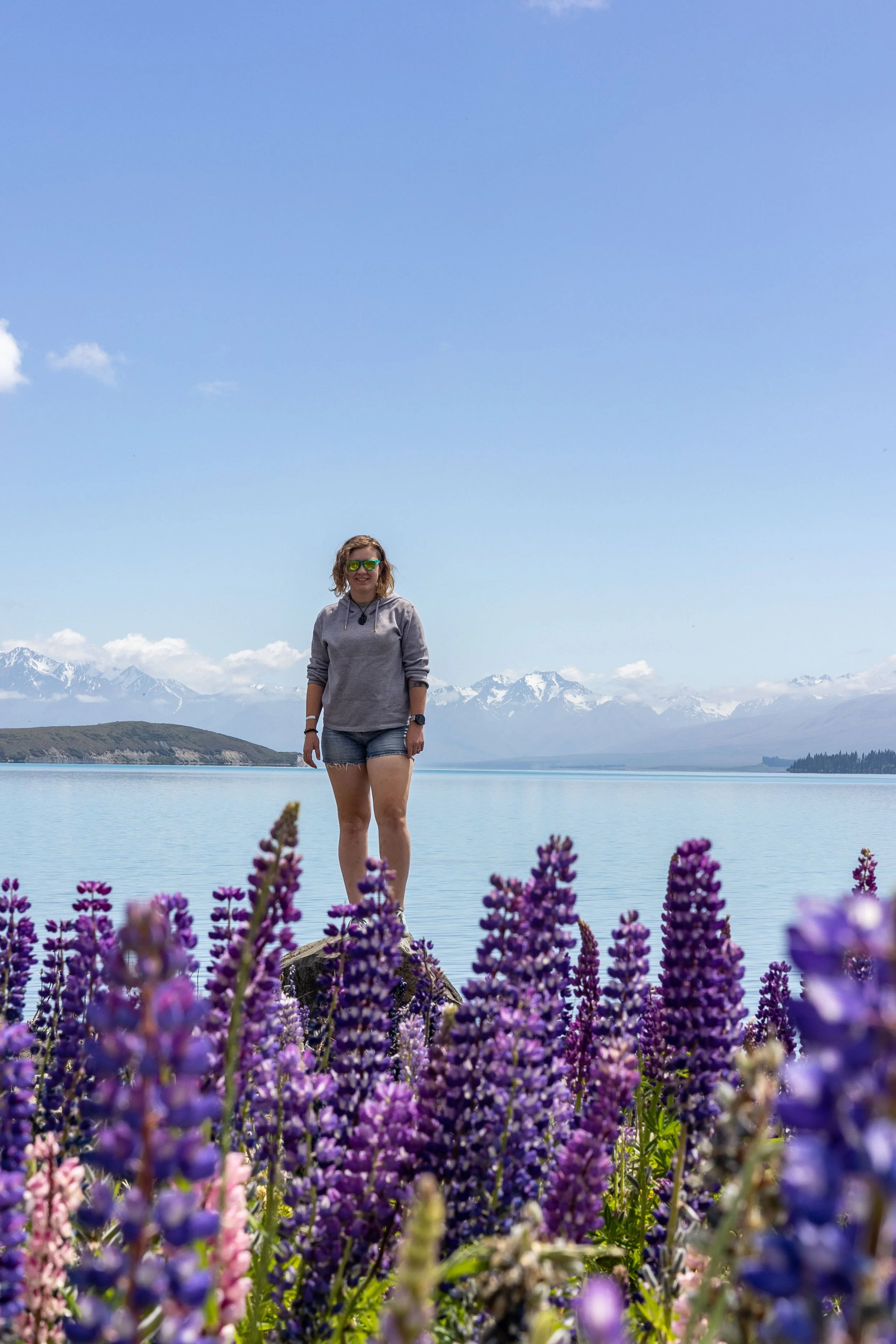 Chelle stands on a rock in front of Lake Tekapo. She is wearing denim shorts and a grey hoodie and green sunglasses. Lupins are blooming bright purple in the foreground. In the background there are snowcapped mountains. Image by Izzi Barton.