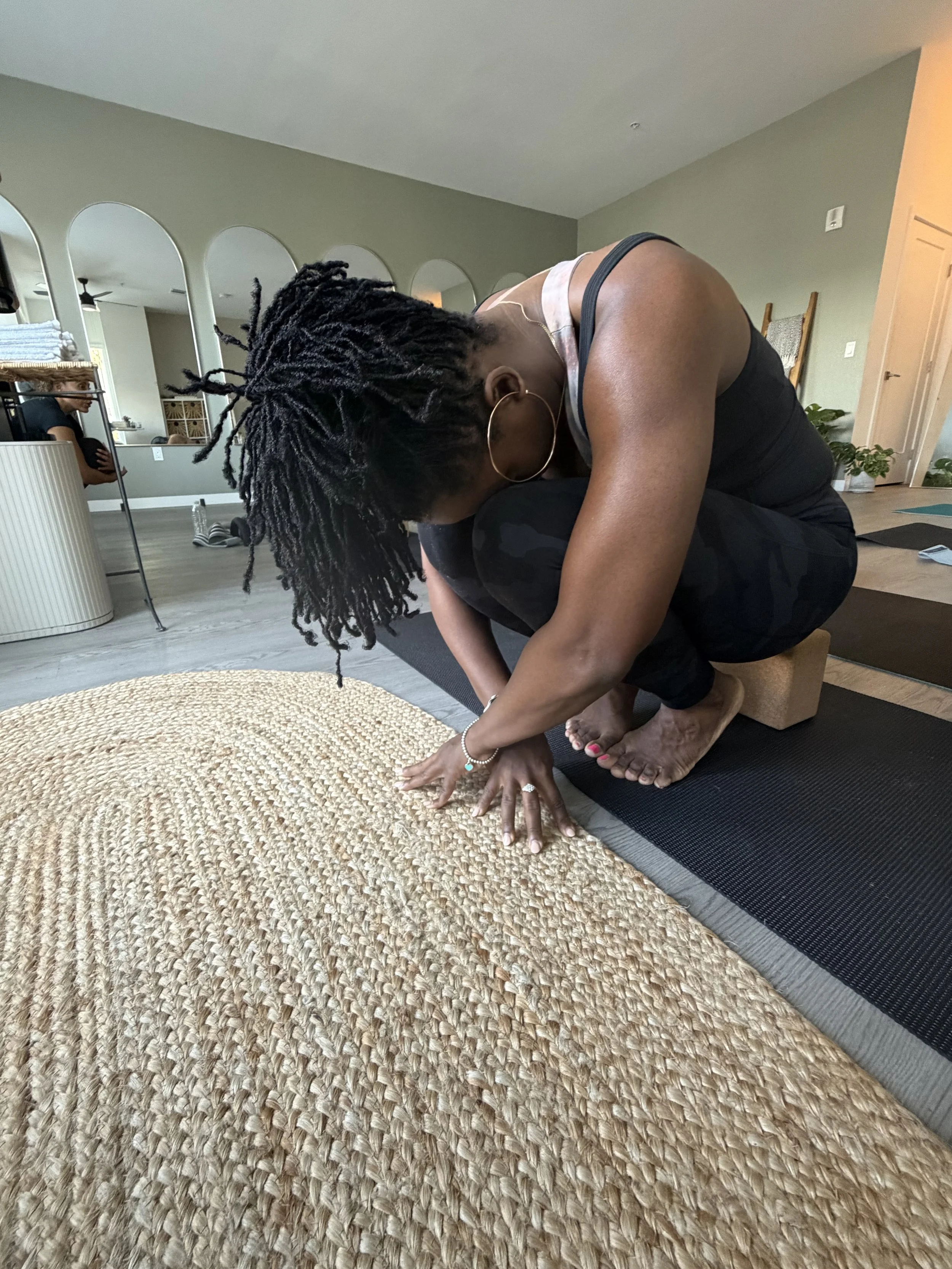 Woman practicing yoga or stretching on a black mat in a living room, with her head bowed down, hands on the floor, and hair tied back. A woven rug is in the foreground, and a small plant is in the background near a chair.