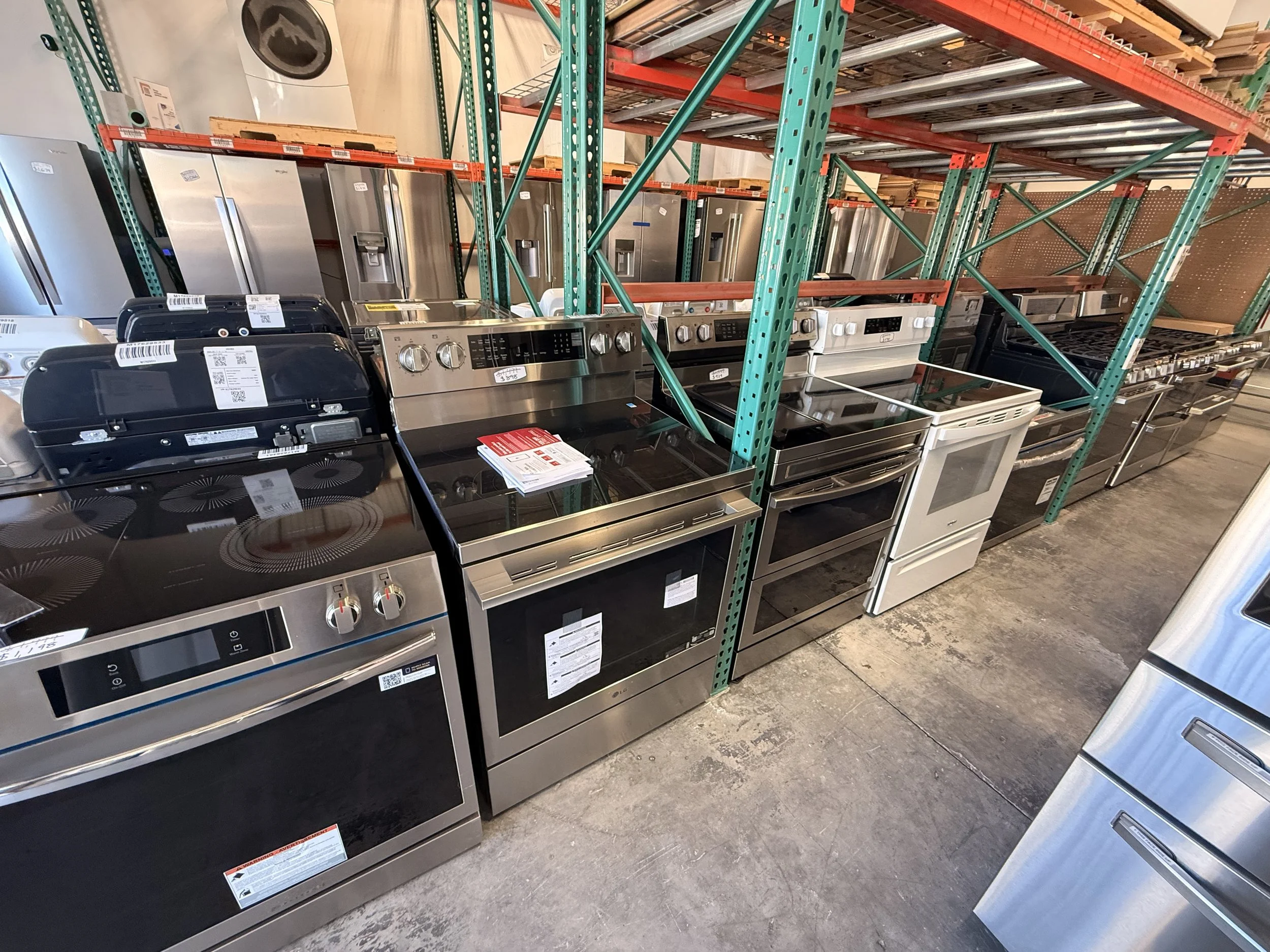Row of stainless steel kitchen appliances, including stove and refrigerators, on store display shelves in a hardware store.