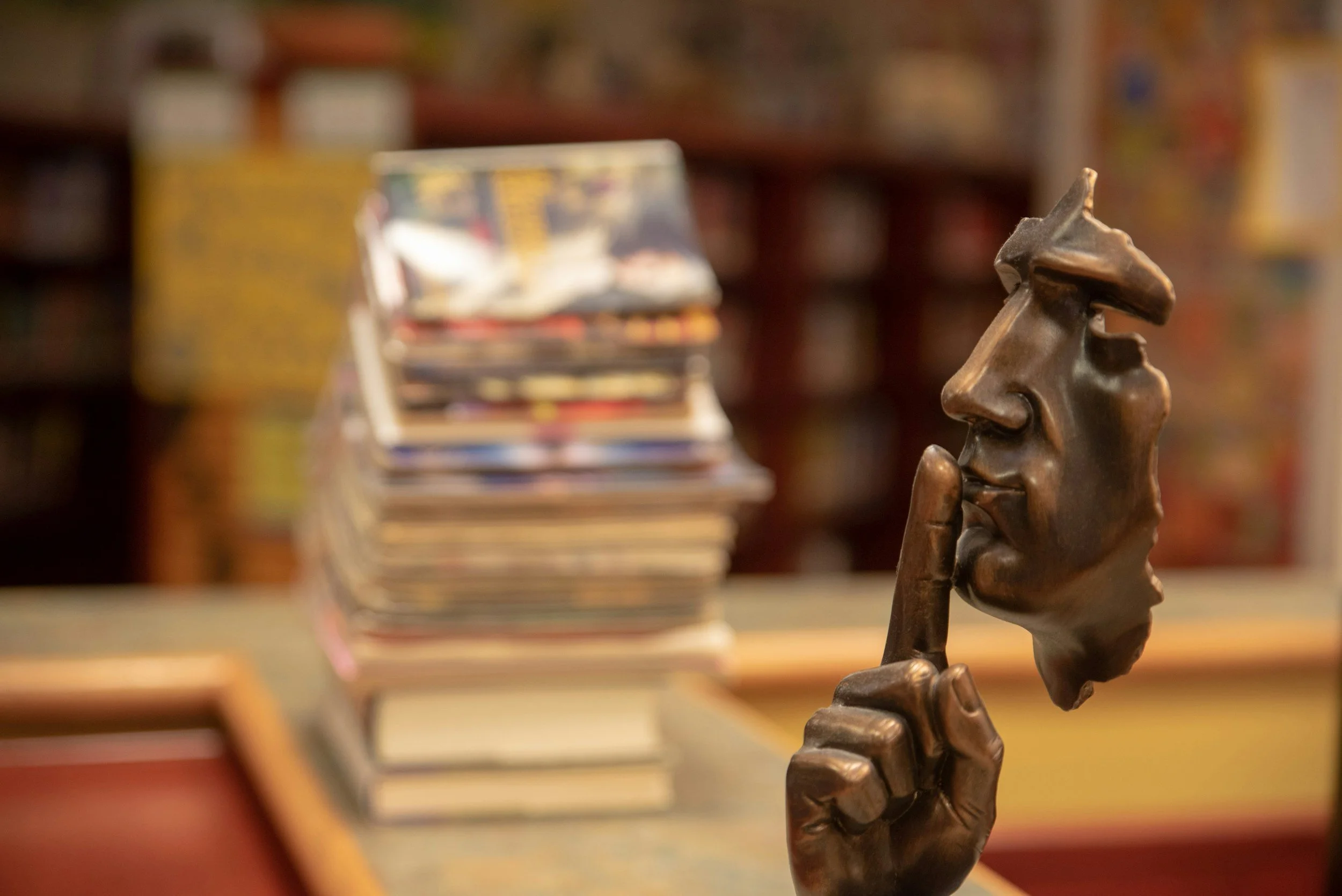 A bronze sculpture of a human face with a finger held up to its lips, indicating silence. In the background, a blurred stack of vinyl records and books in a library or bookstore setting.