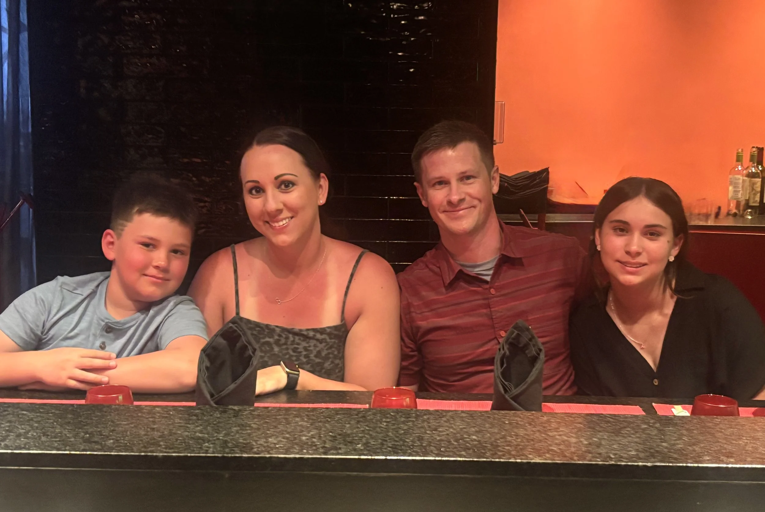Four people sitting at a restaurant table, smiling at the camera. Two women and two boys, with dark and light-colored hair. The background features a dark wall and a section with a reddish-orange hue, with bottles on a shelf.