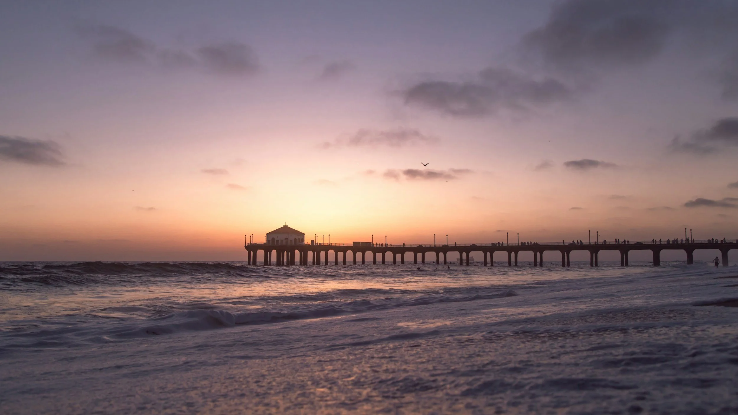 Sunset view of a pier extending into the ocean with small waves and a few people walking on the pier.