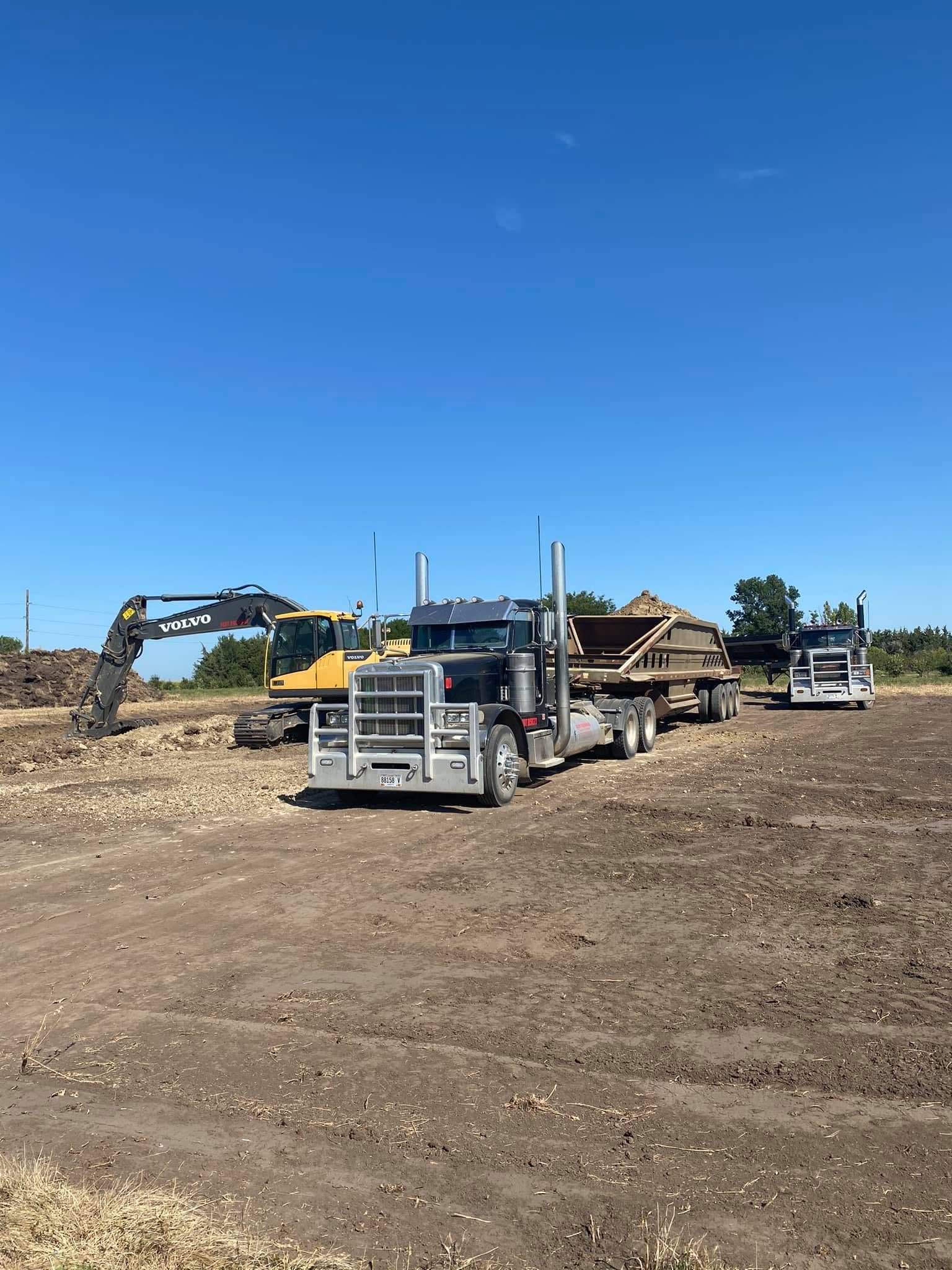 Construction site with a yellow excavator, a semi-truck, and a flatbed trailer on a dirt lot under a clear blue sky.