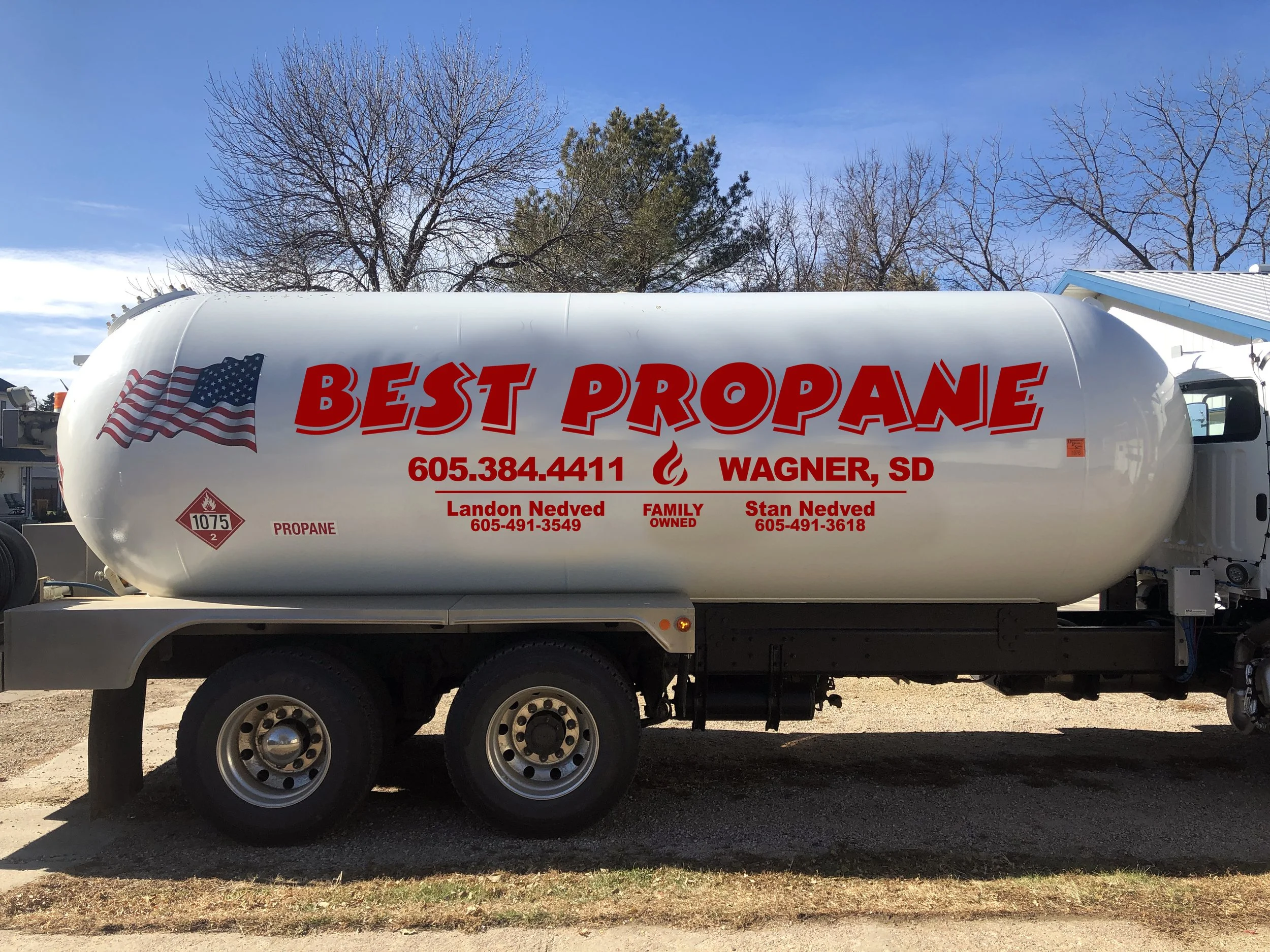A white propane tank on a truck with red and black text, American flag graphic, and contact information, parked outdoors with trees and blue sky in the background.
