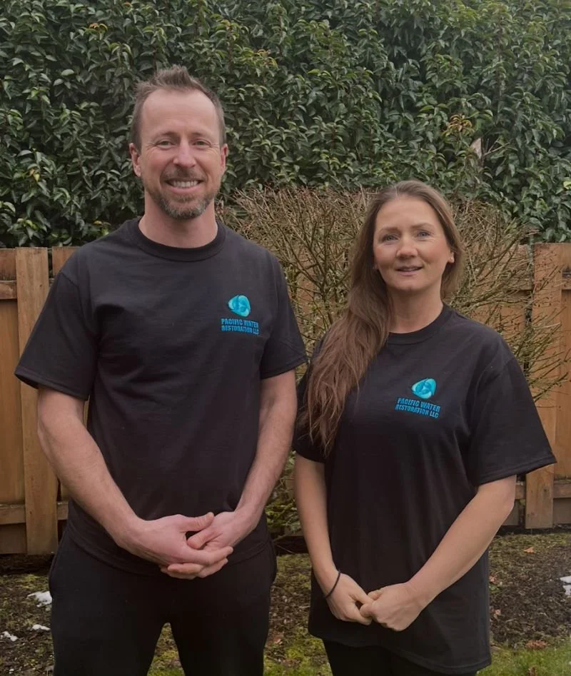A man and woman standing outdoors in front of a wooden fence and greenery, both wearing black t-shirts with a blue logo that says 'Pacific Water Restoration LLC'.