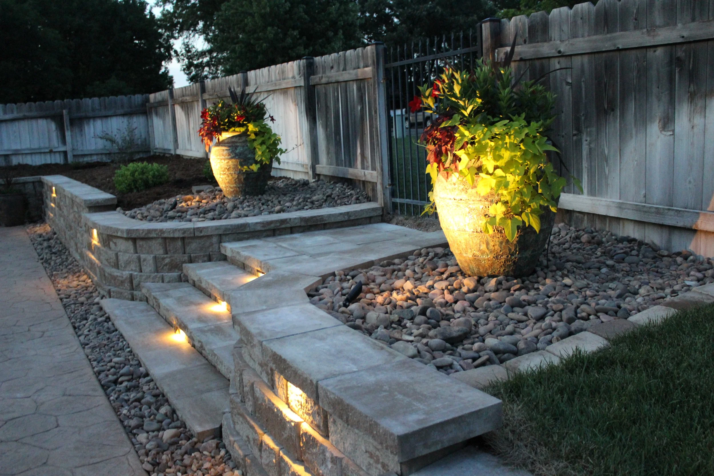 Decorative outdoor garden with two large illuminated flower pots on a gravel bed, a stone staircase with built-in lighting, a wooden fence, a gate, and a paved walkway, during dusk.