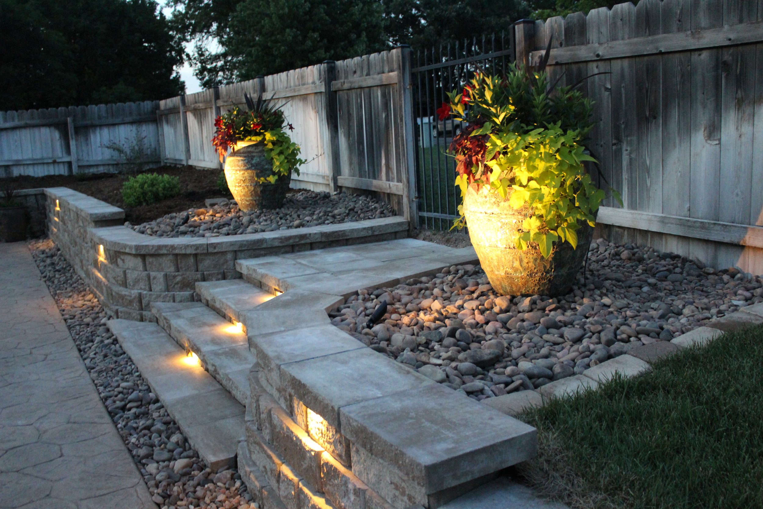 Outdoor garden scene at dusk with two large flower pots illuminated from below, filled with colorful plants. The garden features a curved stone retaining wall with built-in lighting, stone steps, and a gravel ground cover, enclosed by a wooden fence.