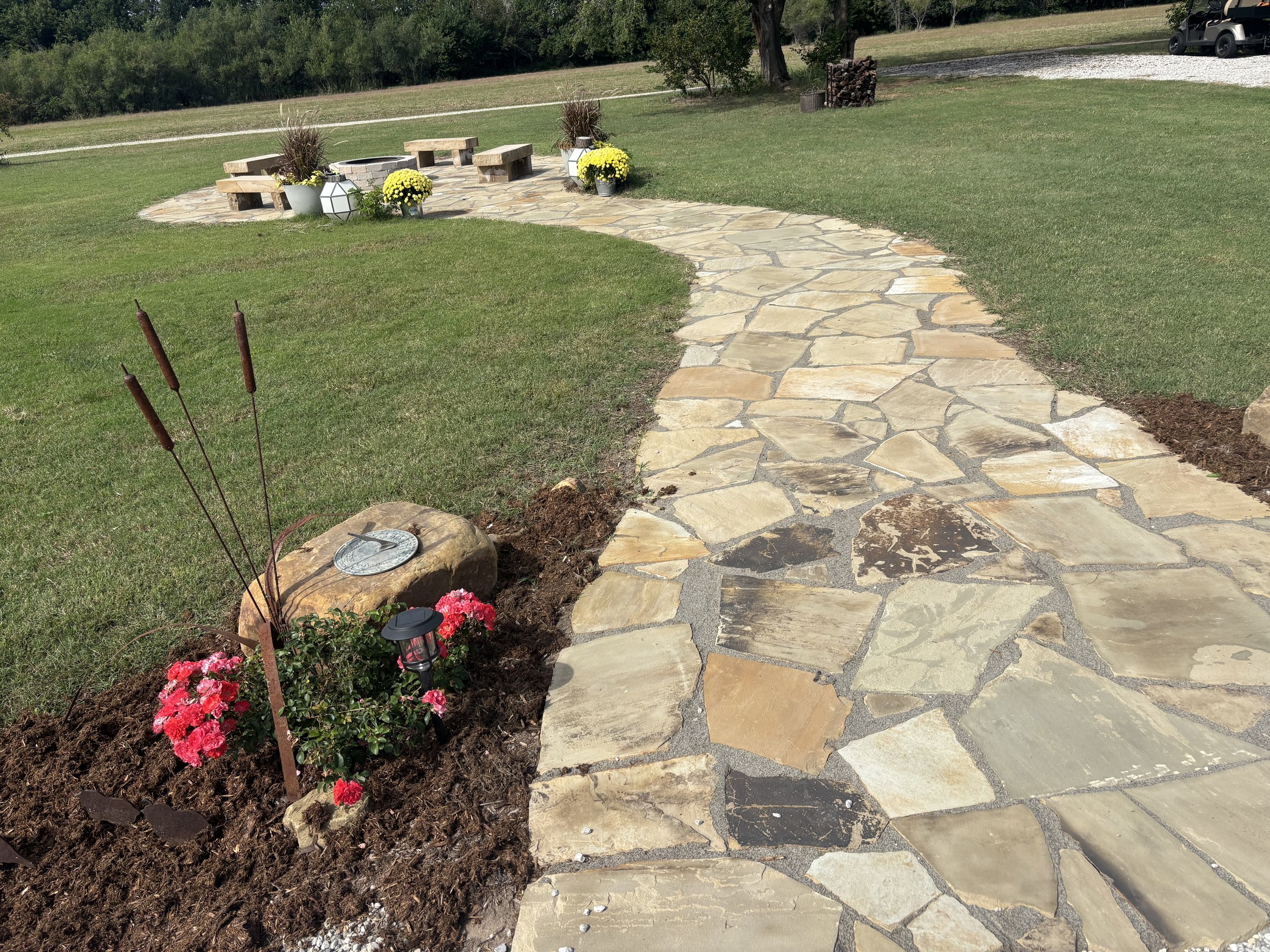 A curved stone pathway leading to a backyard patio with flower pots and wooden benches. Front yard flower bed with red flowers and garden decorations including a large rock, a clock, and stake plants. Green lawn, trees, gravel driveway, and a vehicle in the background.