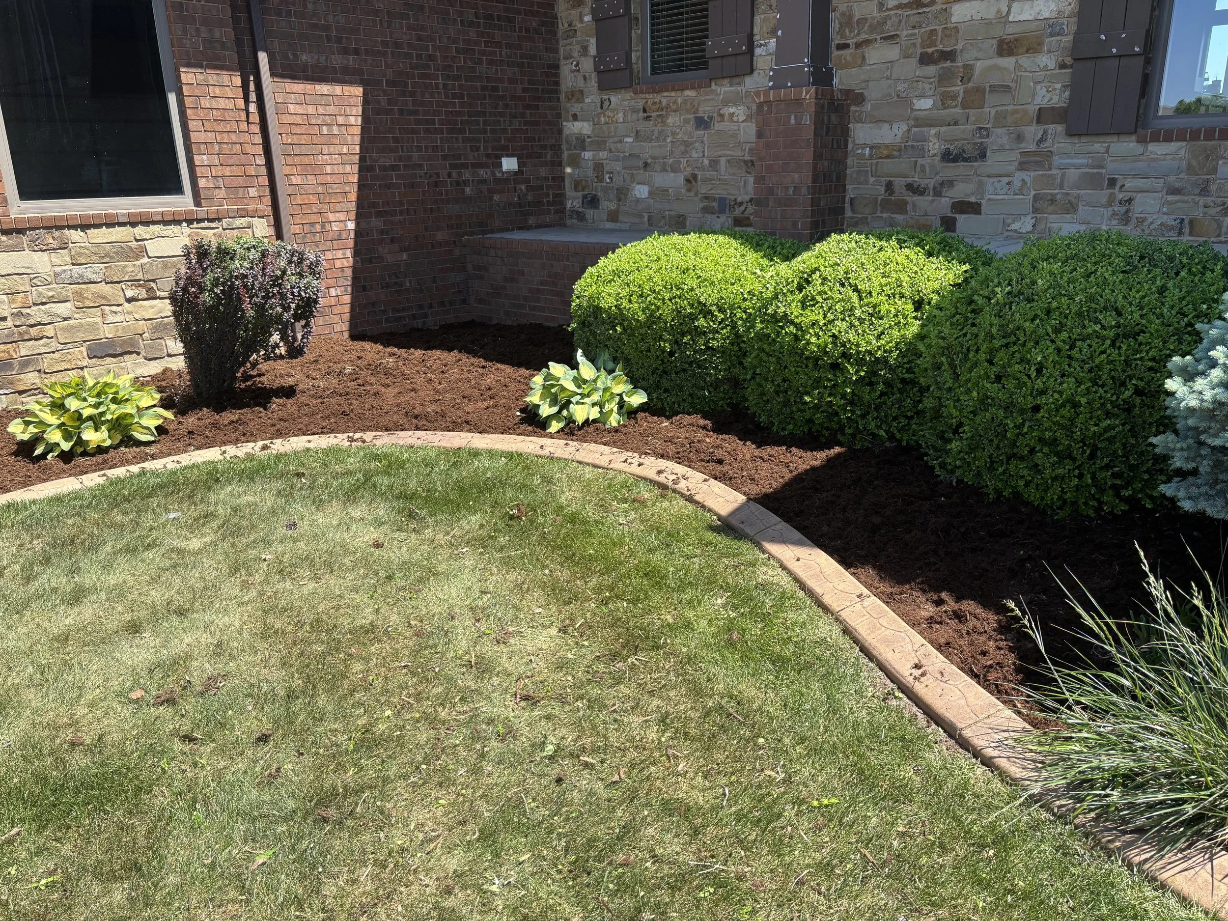 A landscaped front yard with green grass, a curved brick edging, and various bushes and plants against a house with brick and stone exterior.