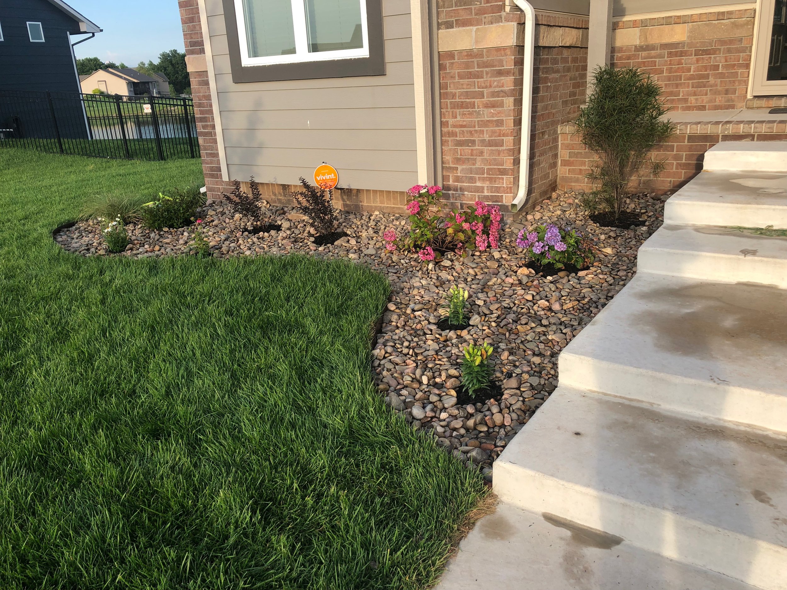 Front yard garden with a well-maintained green lawn, colorful flowers, small shrubs, and a brick house with concrete steps.