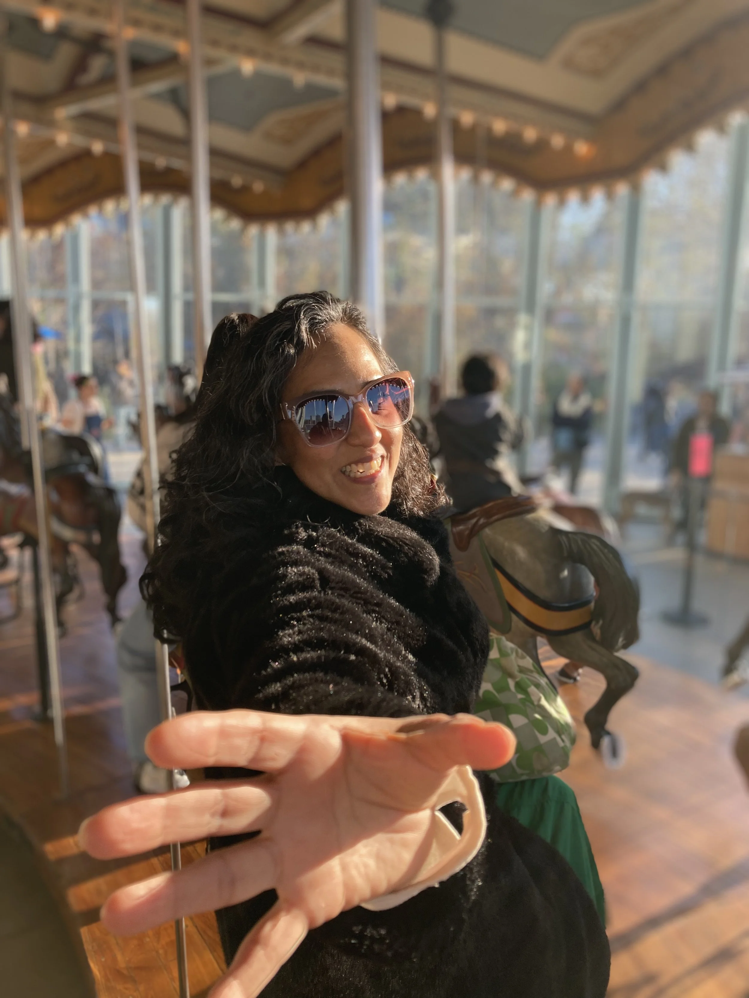 Woman with dark, curly hair, wearing sunglasses and a black fur coat, smiling and reaching out with her hand inside a carousel ride area.