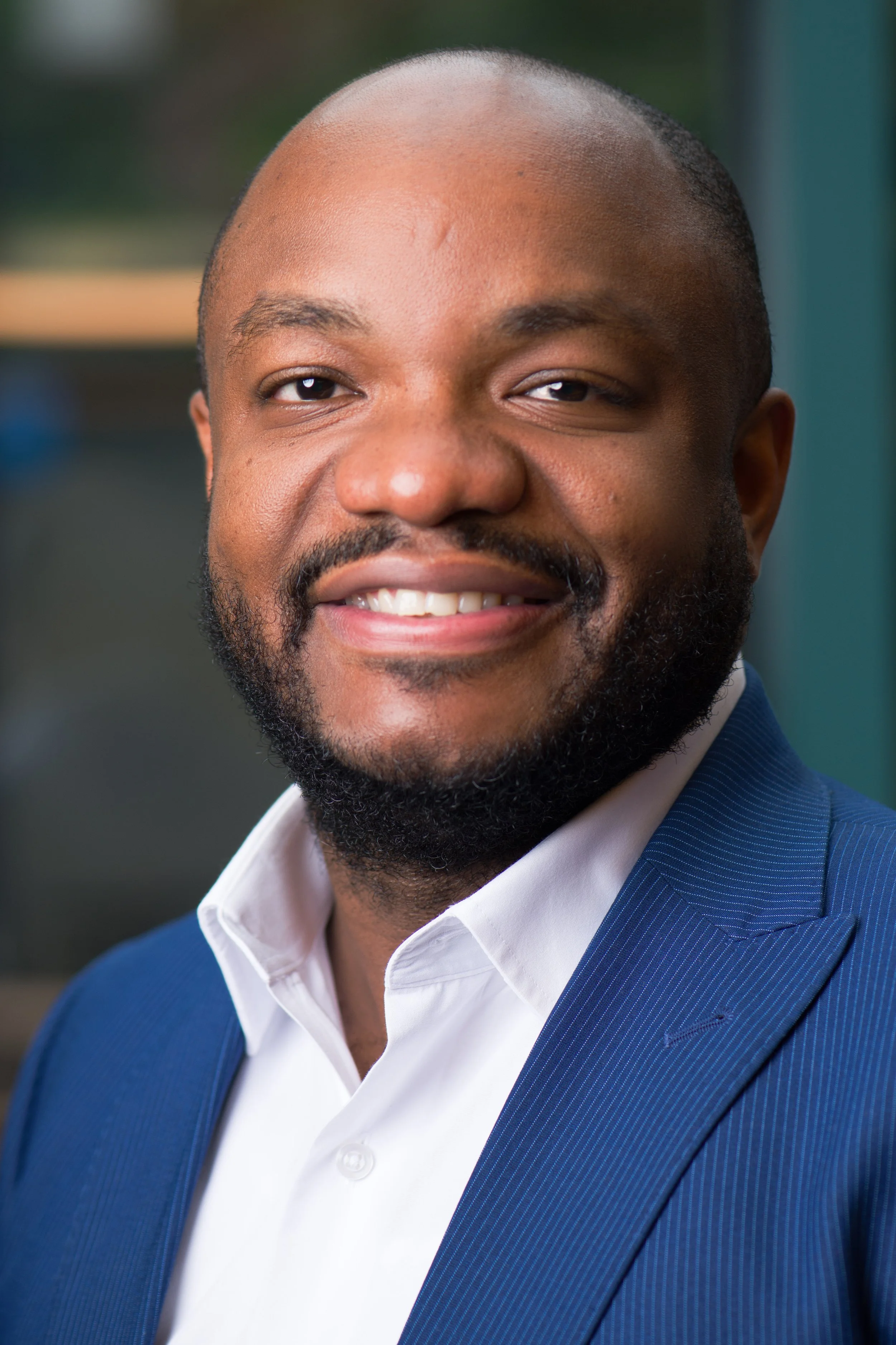 Close-up portrait of a smiling man with a beard, wearing a blue blazer and white shirt, with a blurred outdoor background.